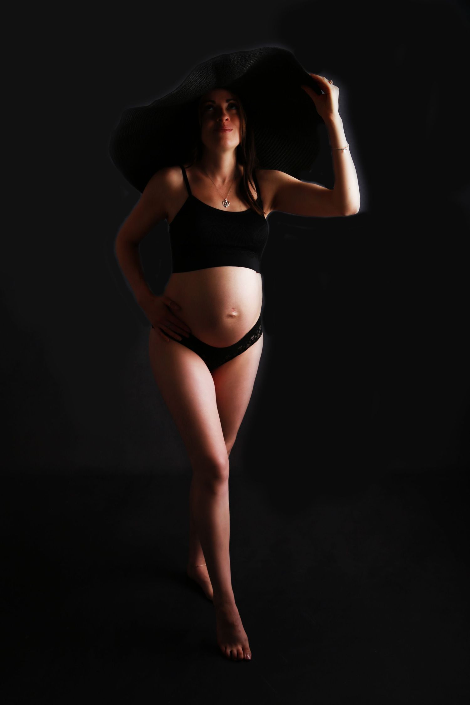 Pregnant woman in black lingerie and wide-brim hat posing in dramatic dark studio.