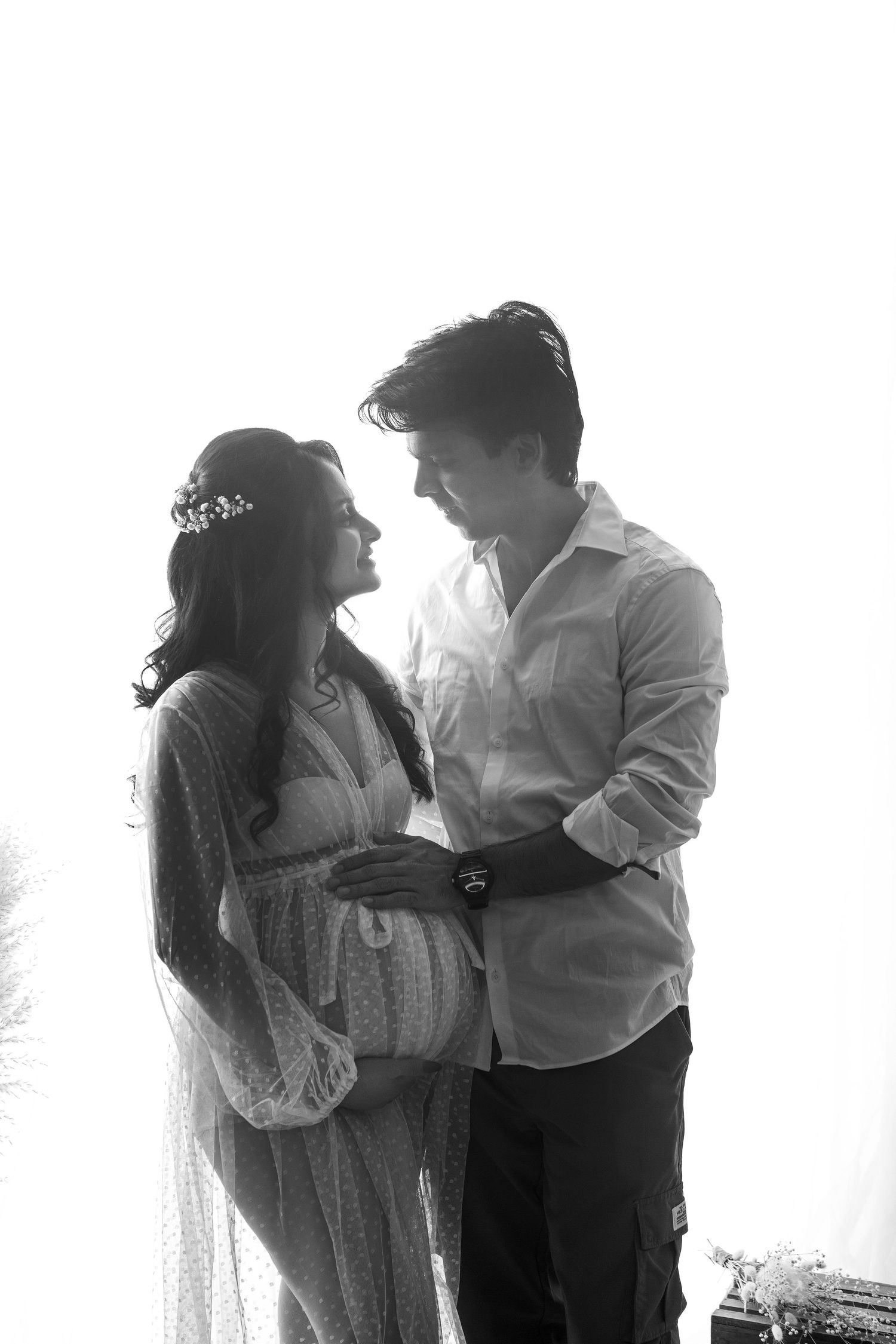 Black and white photo of smiling pregnant couple embracing, woman wearing crown headpiece in bright studio.