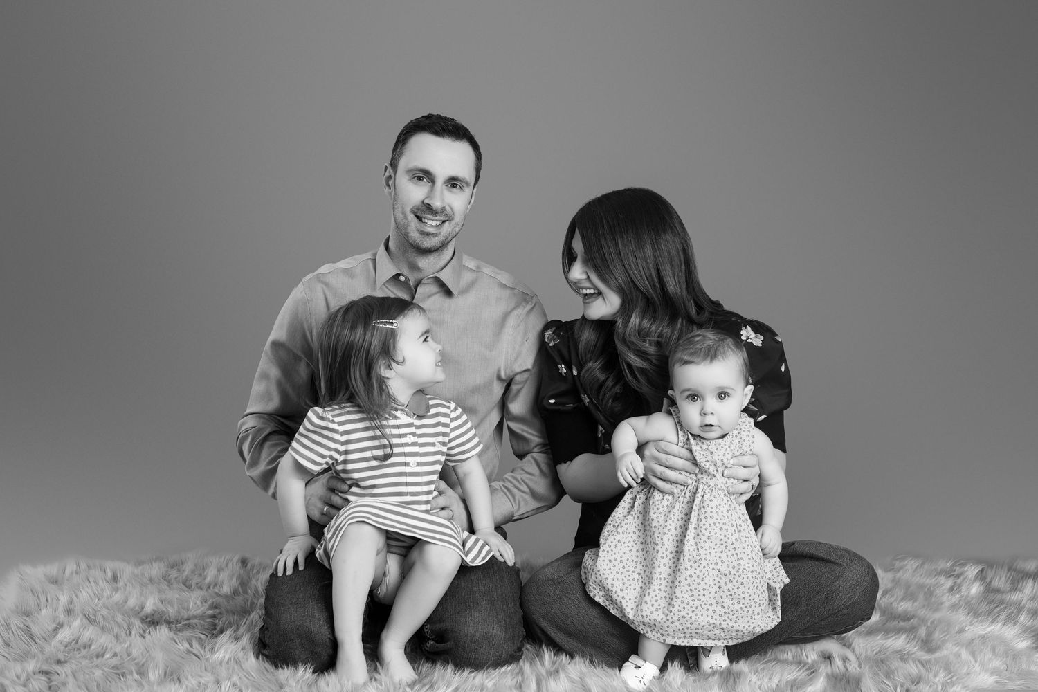 Black and white family portrait of parents with two young daughters sitting on a fluffy rug in a studio.