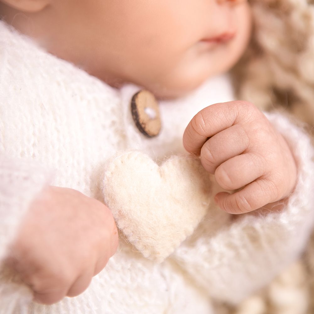 A baby in a white knit sweater holds a small fluffy heart-shaped toy in their tiny hands.