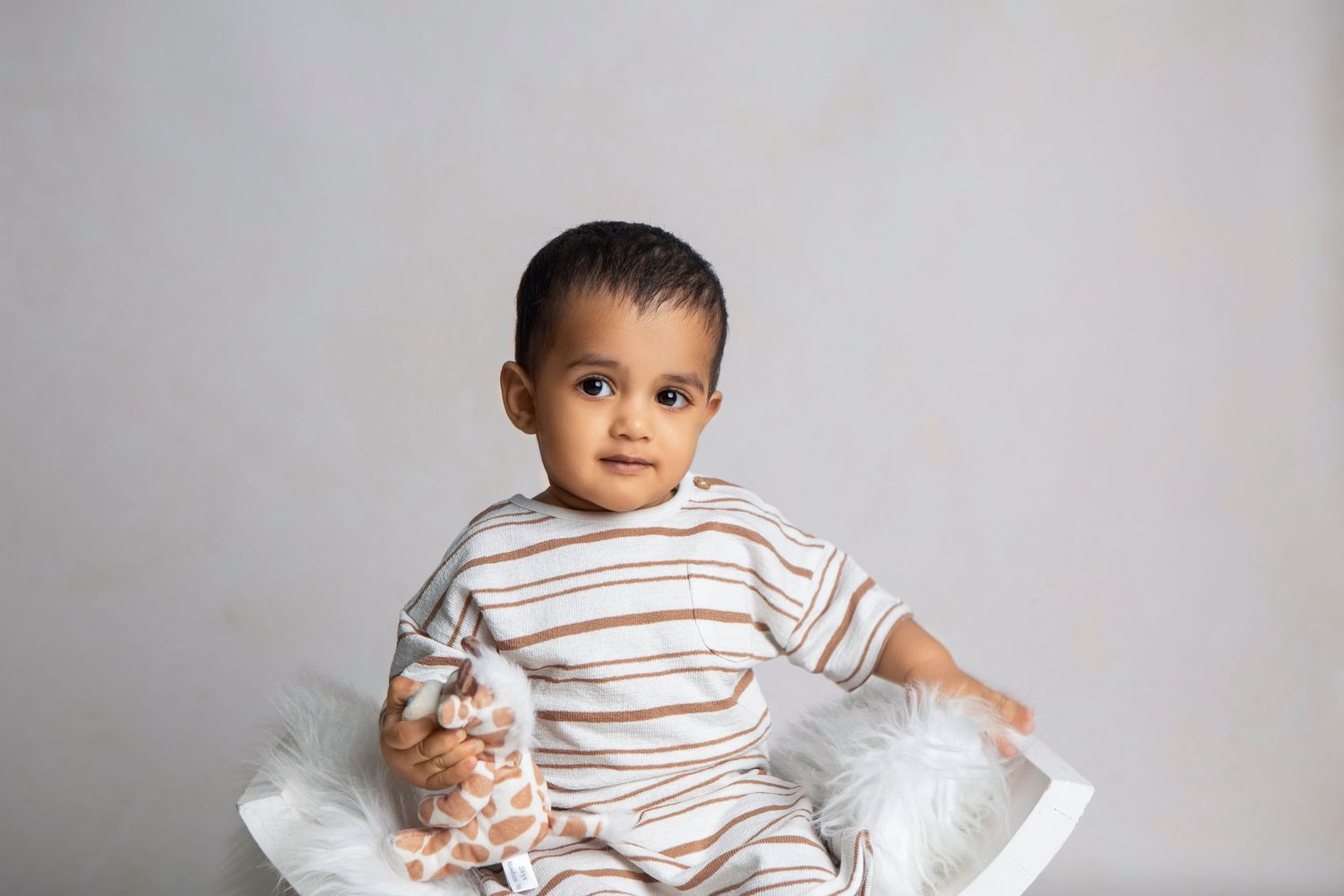 Adorable baby boy in striped pajamas sitting in a white chair holding a jar of treats against a gray background.
