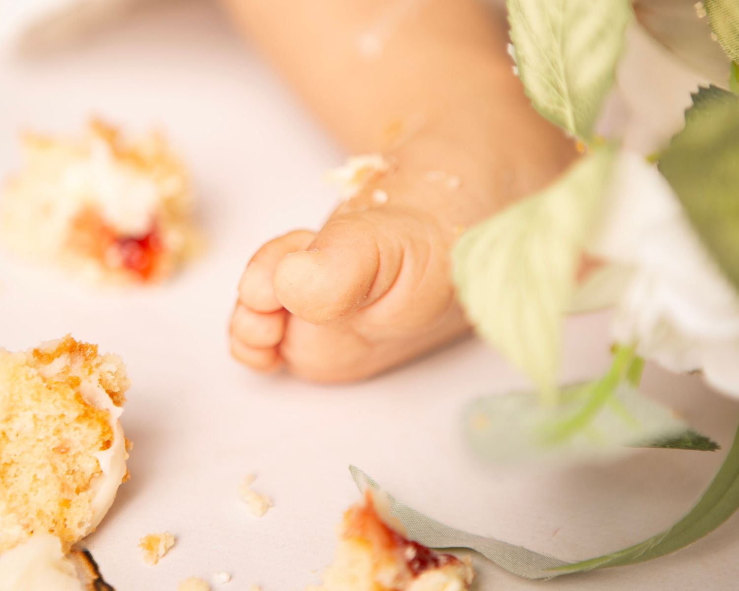 Baby fist next to crumbled birthday smash cake pieces and white flower on white surface.