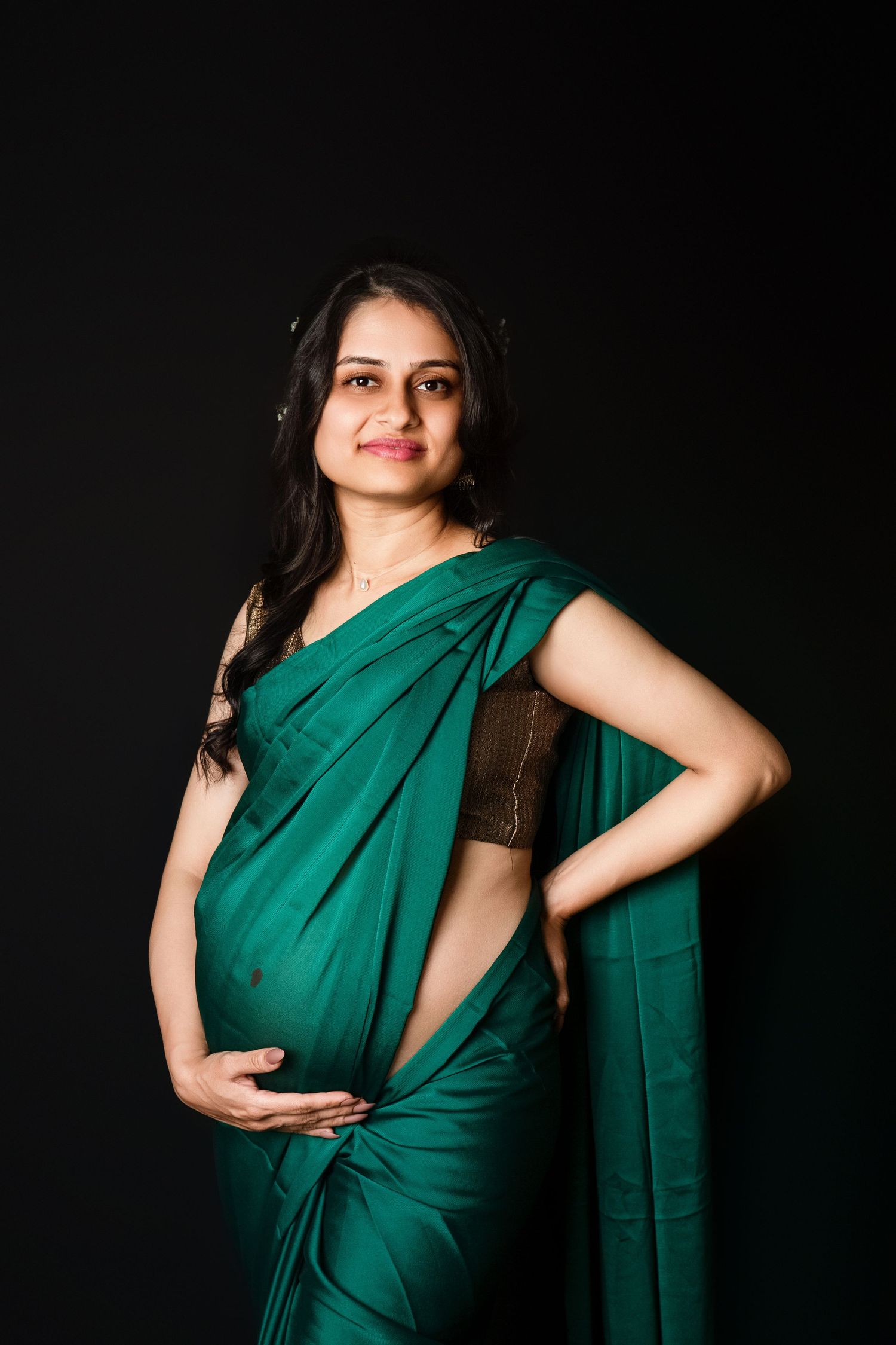 Smiling pregnant Indian woman in teal green saree with hand on hip against black background.