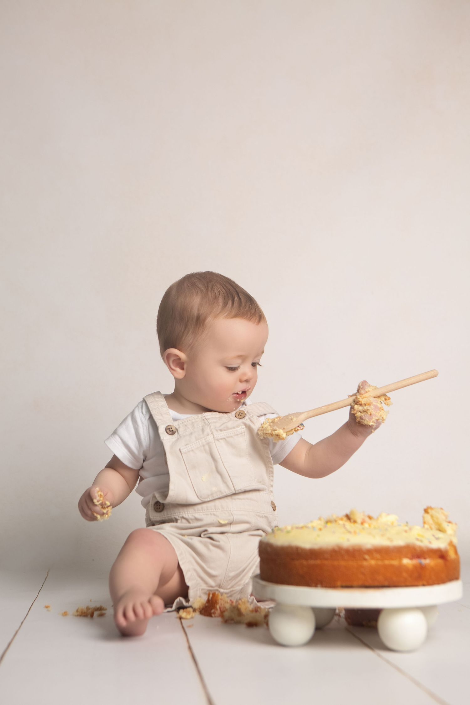 Cute baby in beige overalls smashing a first birthday cake with a wooden spoon on a white floor.