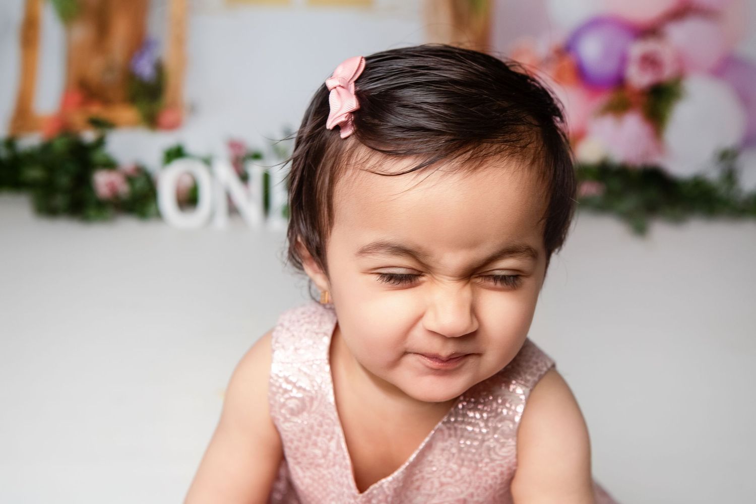 Cute baby girl in pink dress squinting and smiling during first birthday cake smash photo session.