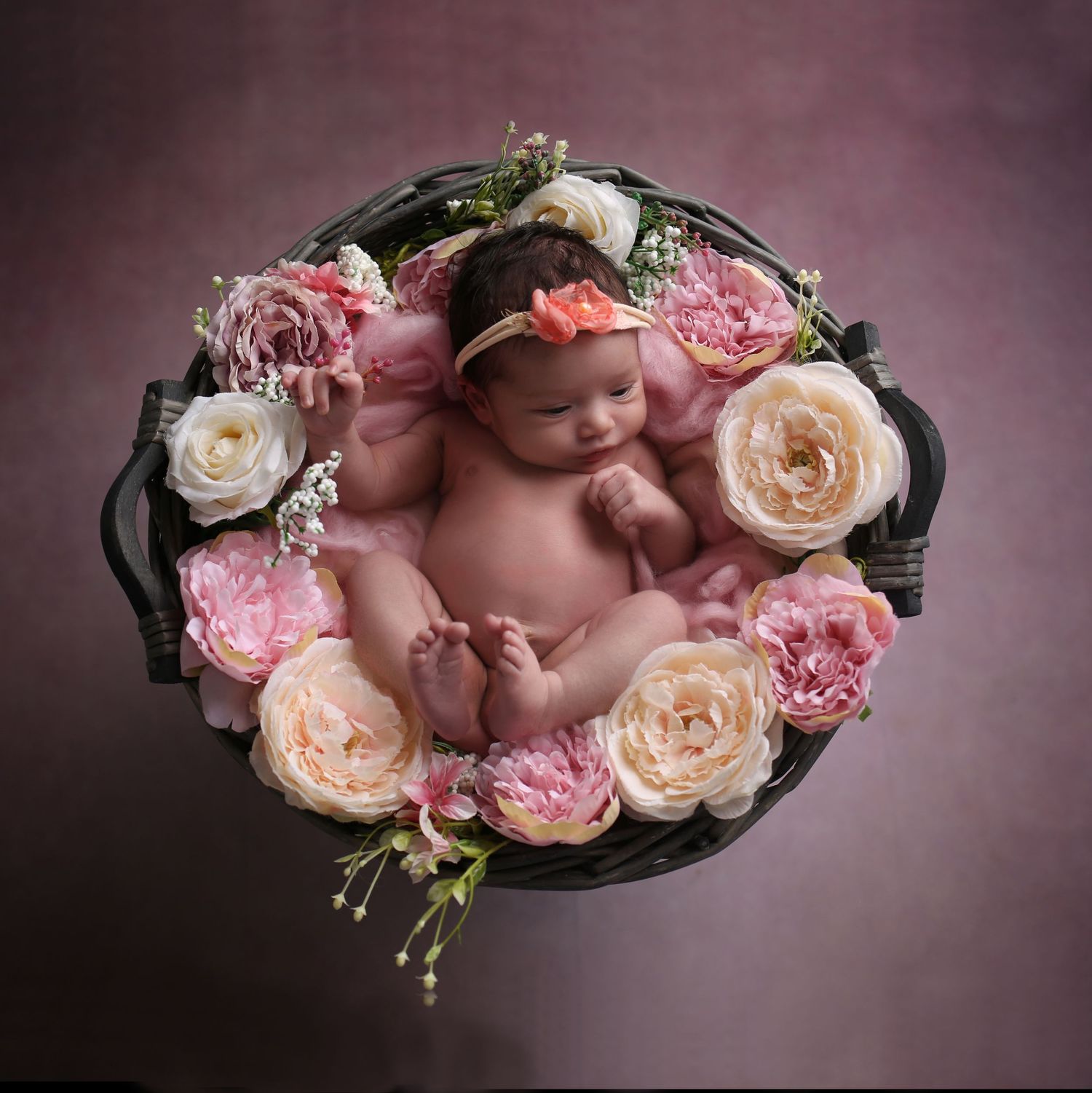 Newborn baby sleeping in a wicker basket surrounded by pink, peach, and white flowers, wearing a floral headband.