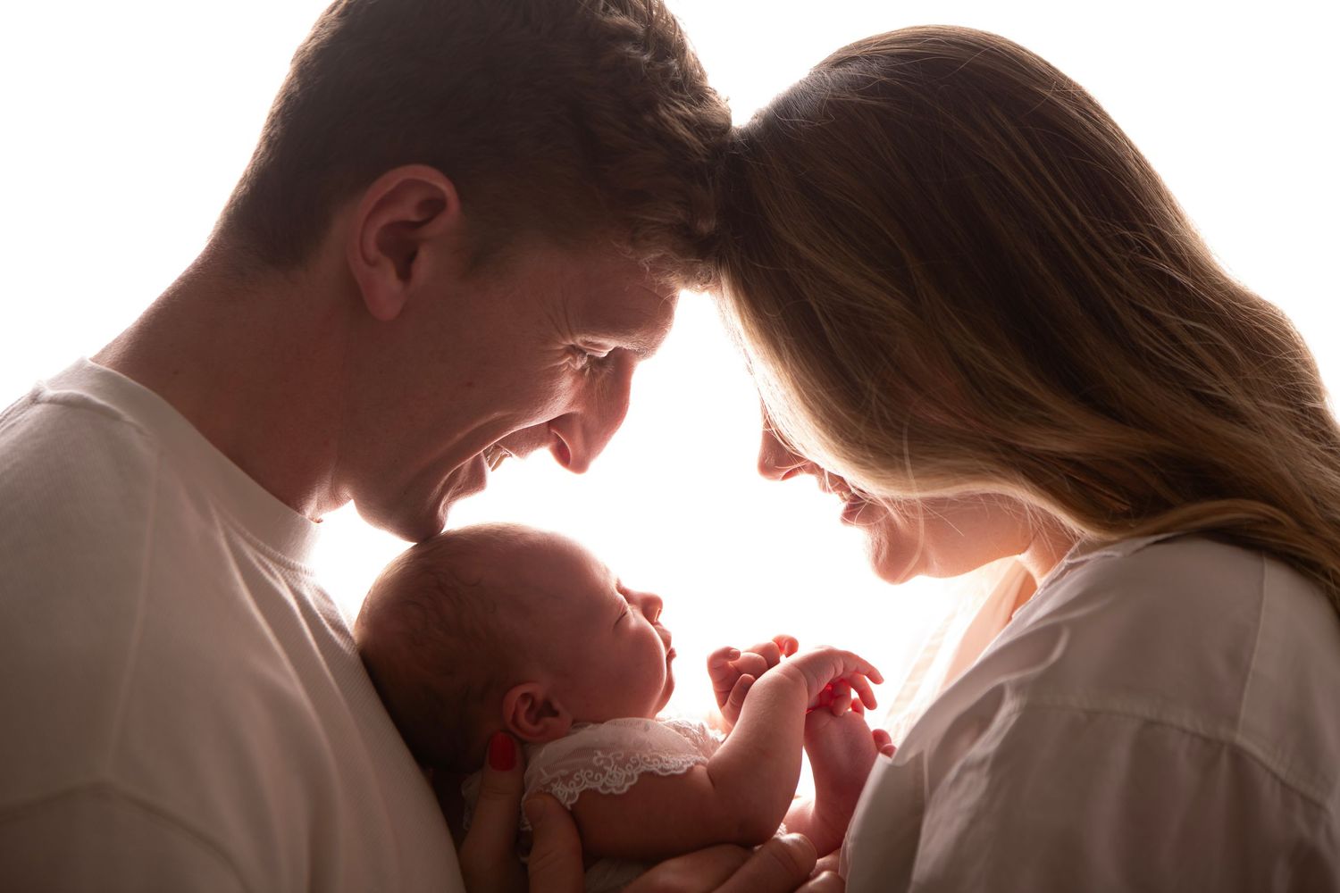 Happy parents smiling at their newborn baby in a bright, backlit family portrait.