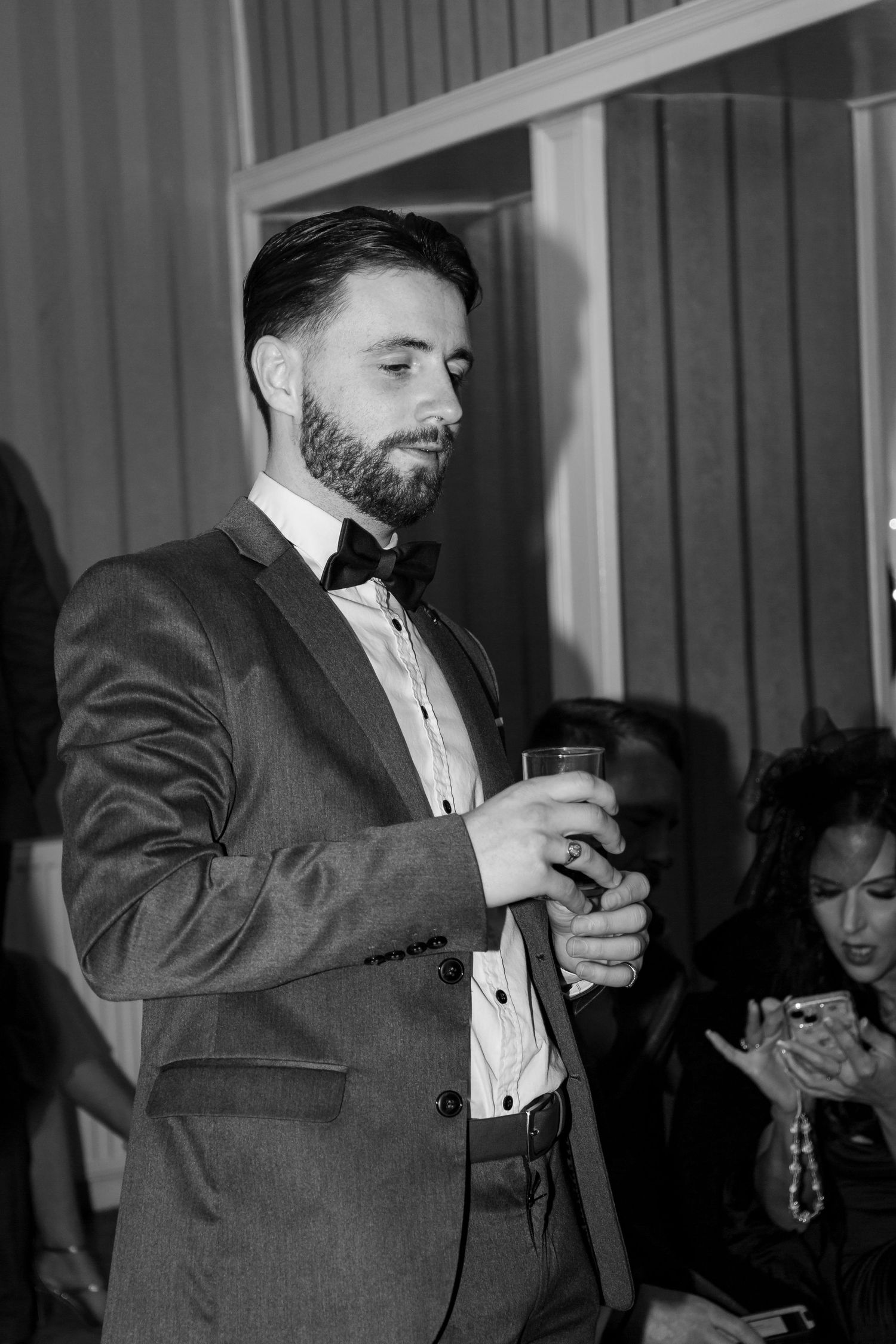 Bearded man in gray suit and bow tie holding a drink glass at a formal evening event.