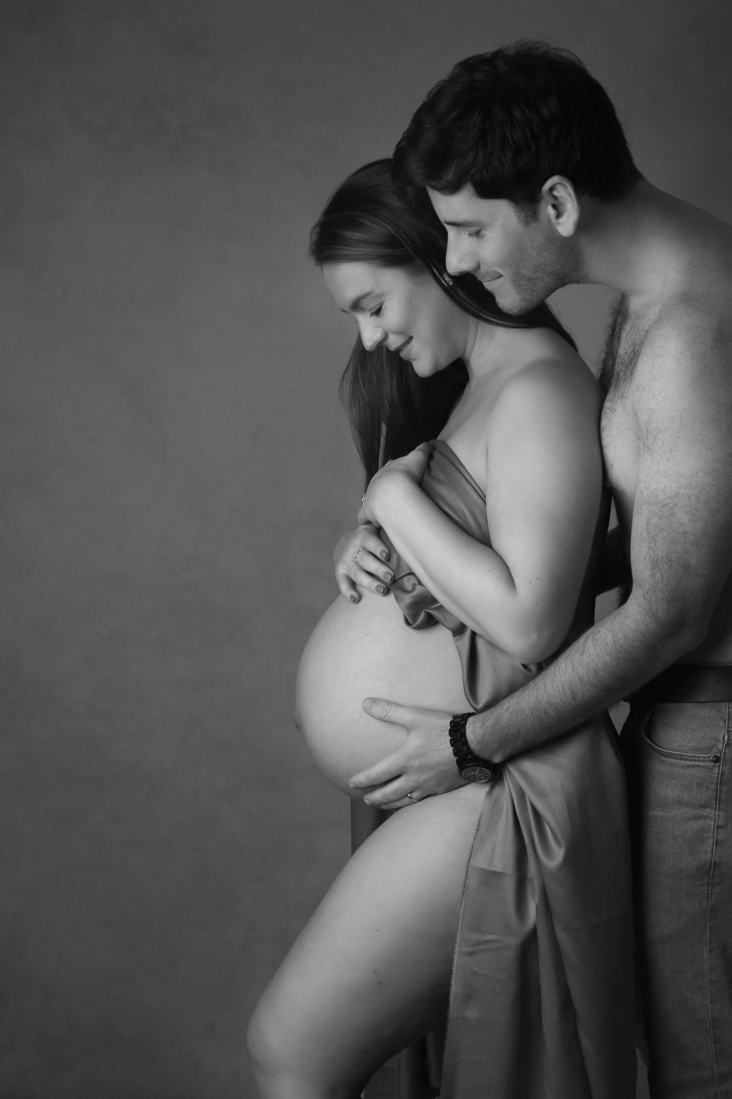 Black and white maternity photo of pregnant woman and partner tenderly embracing, cradling baby bump in studio.
