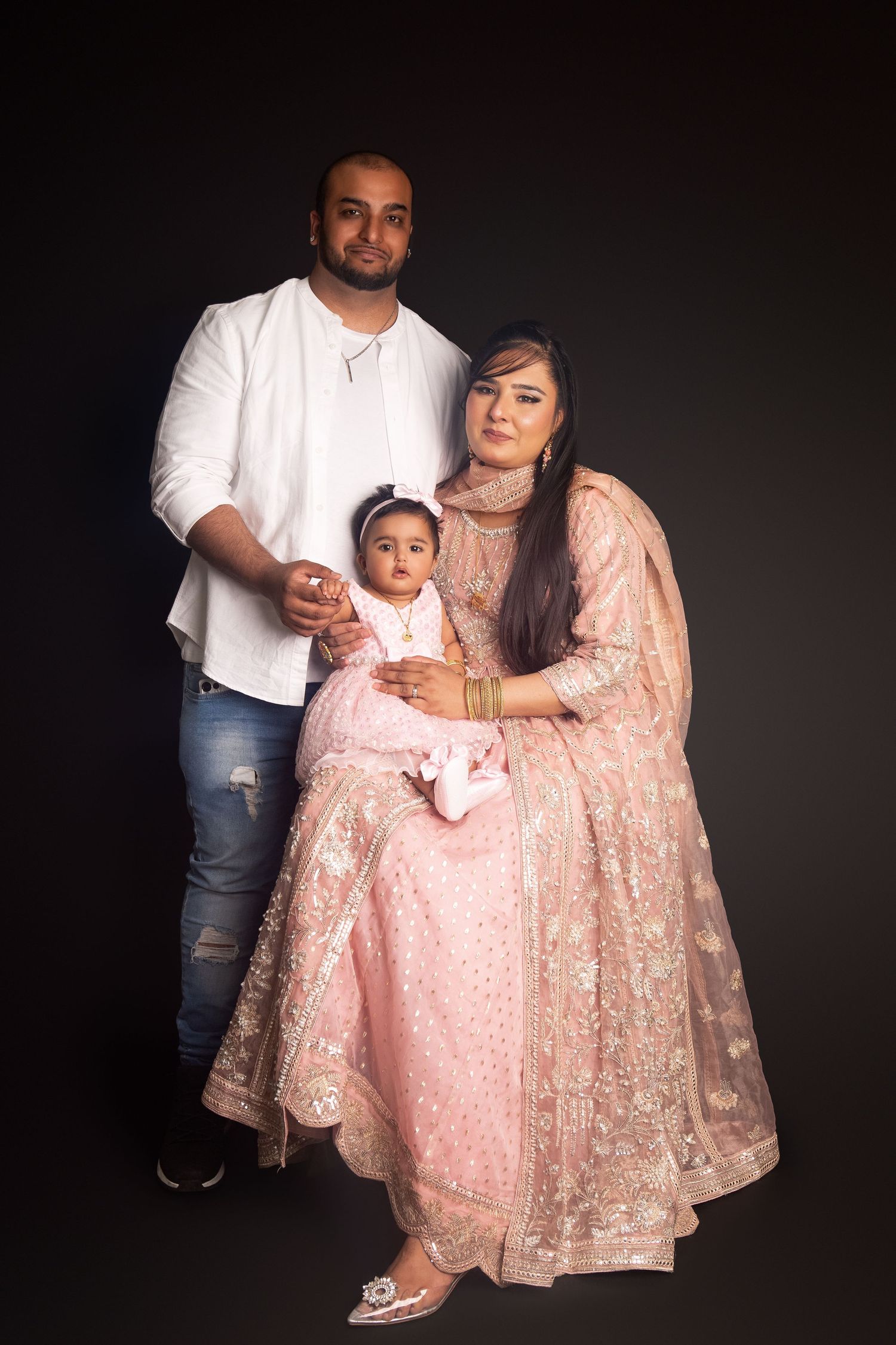 A South Asian family posing in studio, man in white shirt, woman in pink lehenga holding a baby girl.