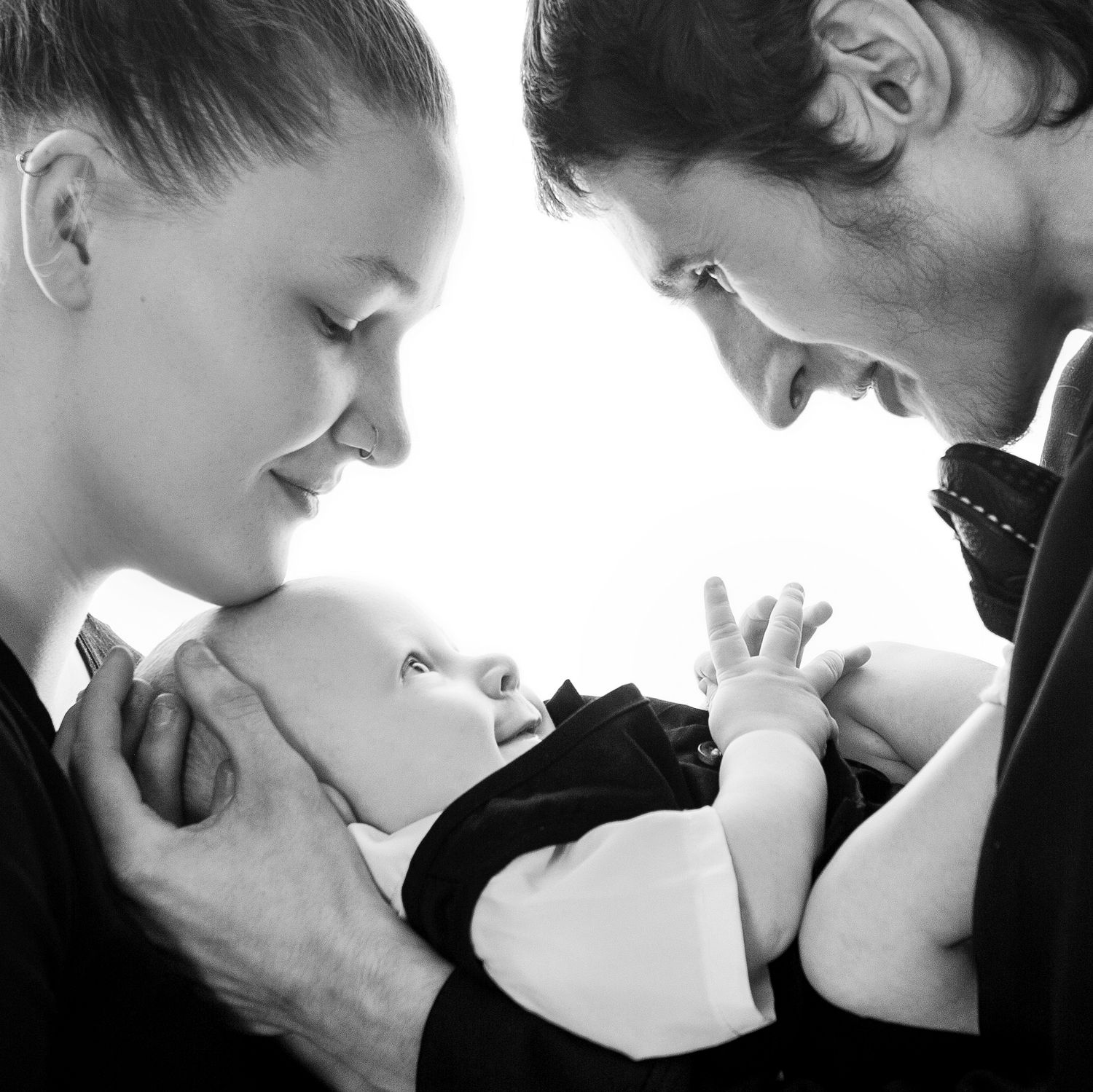 Black and white close-up of loving parents gazing down at alert newborn baby held between them.