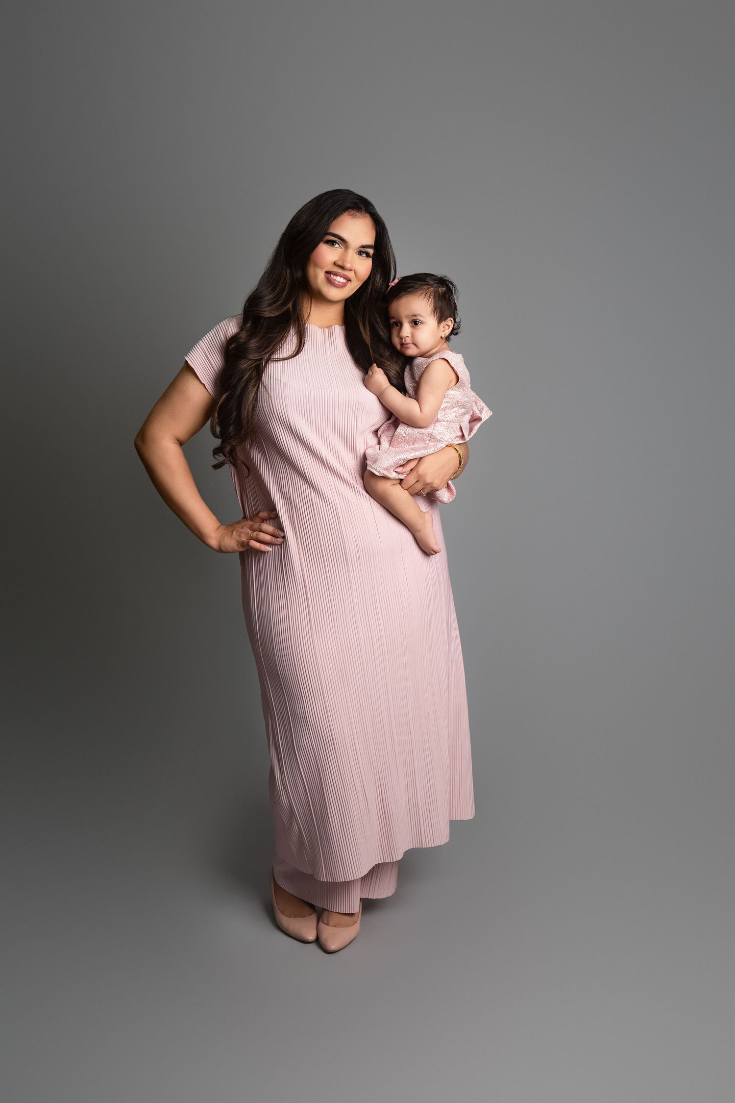 A woman in a pink maxi dress holds her baby girl, both dressed in matching pink outfits, posing for a studio portrait.