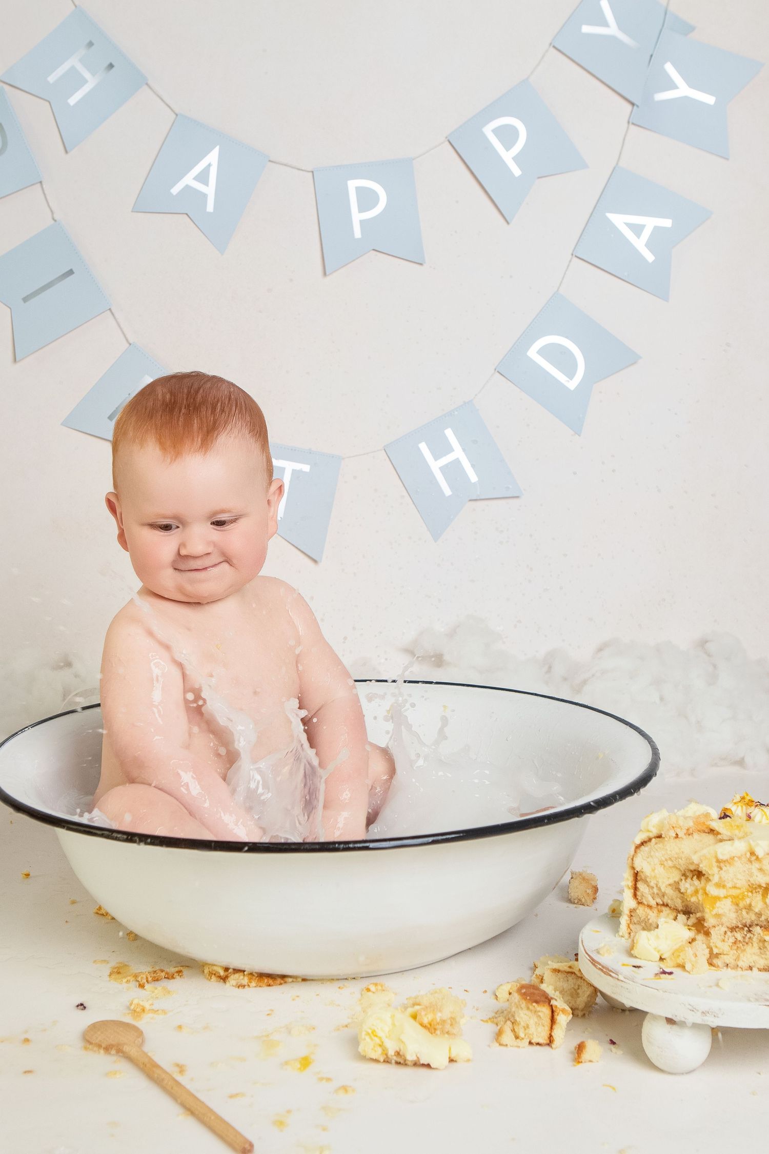 Baby sitting in a white basin during a first birthday cake smash session with a Happy Birthday banner.