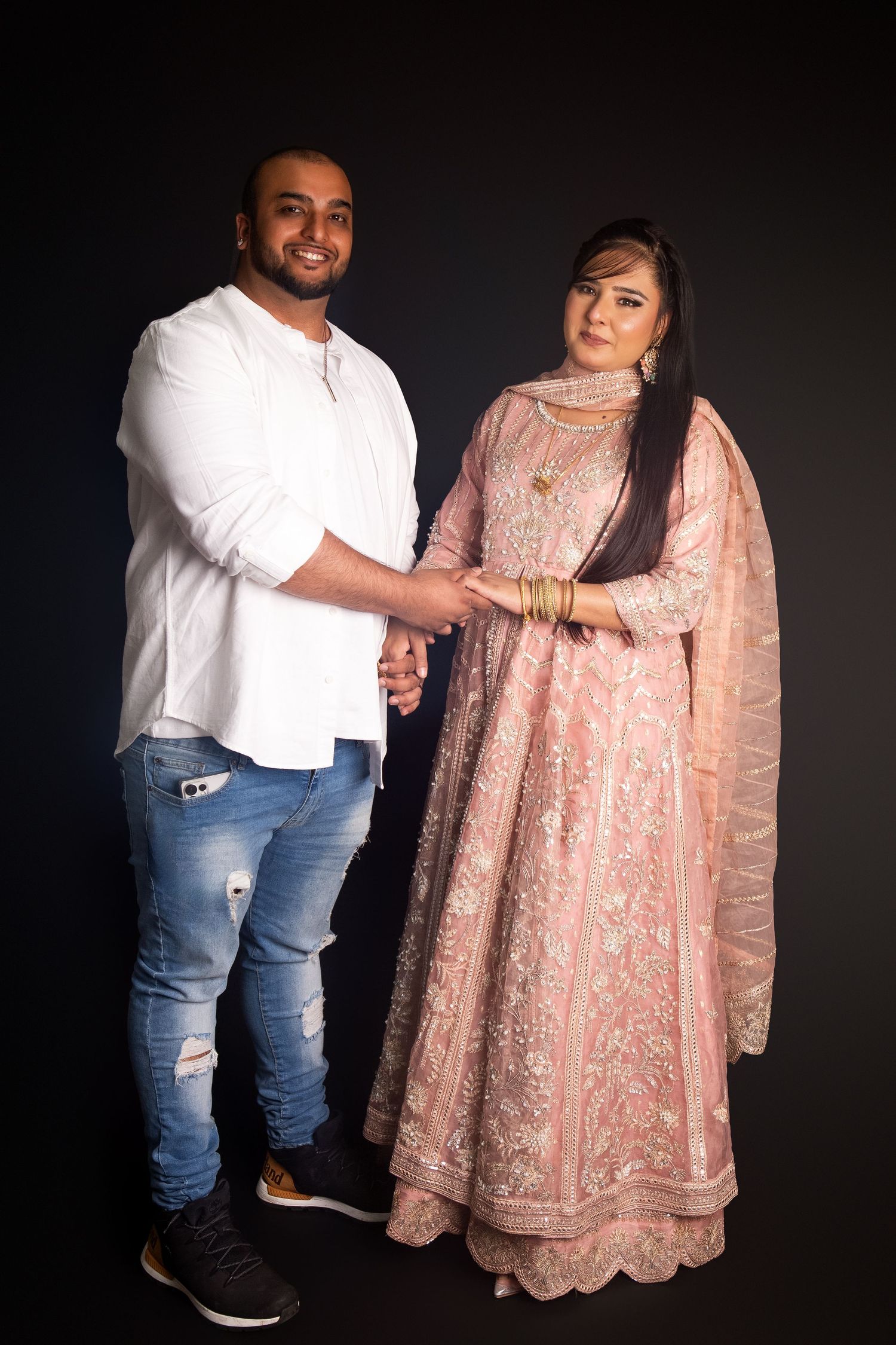A couple in traditional and casual attire hold hands against a dark background, the woman wearing a pink embroidered anarkali suit.