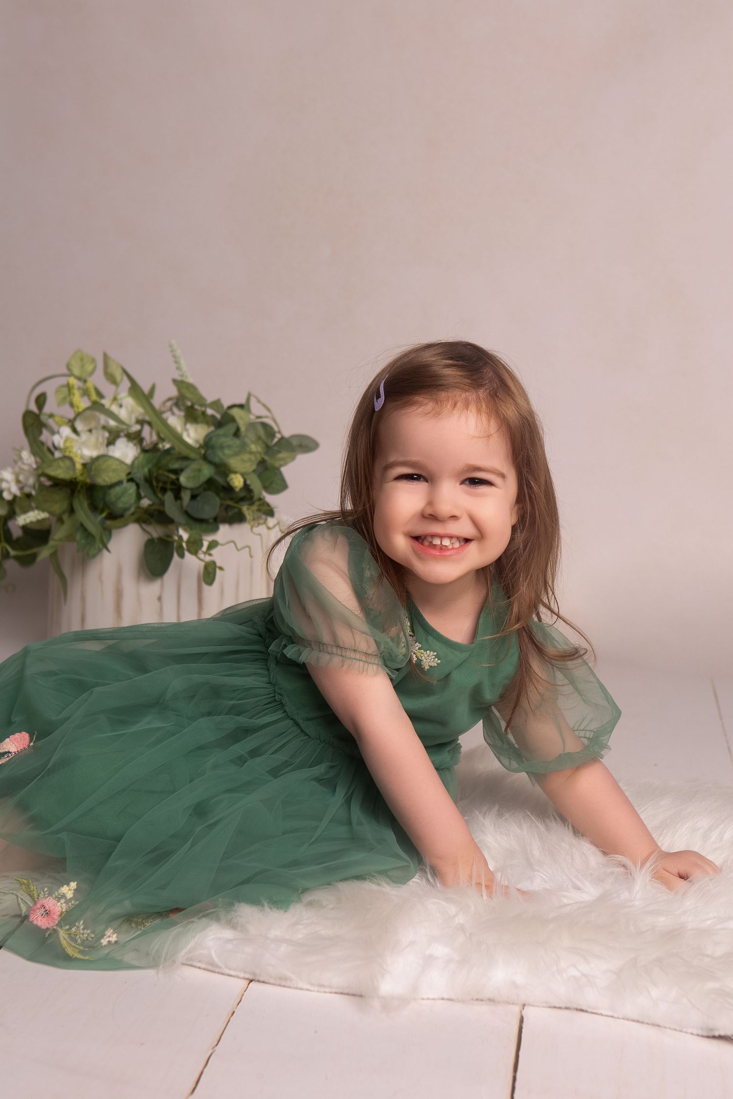 A smiling toddler girl in a green tulle dress sits on a white fur rug beside white flowers in a studio portrait.