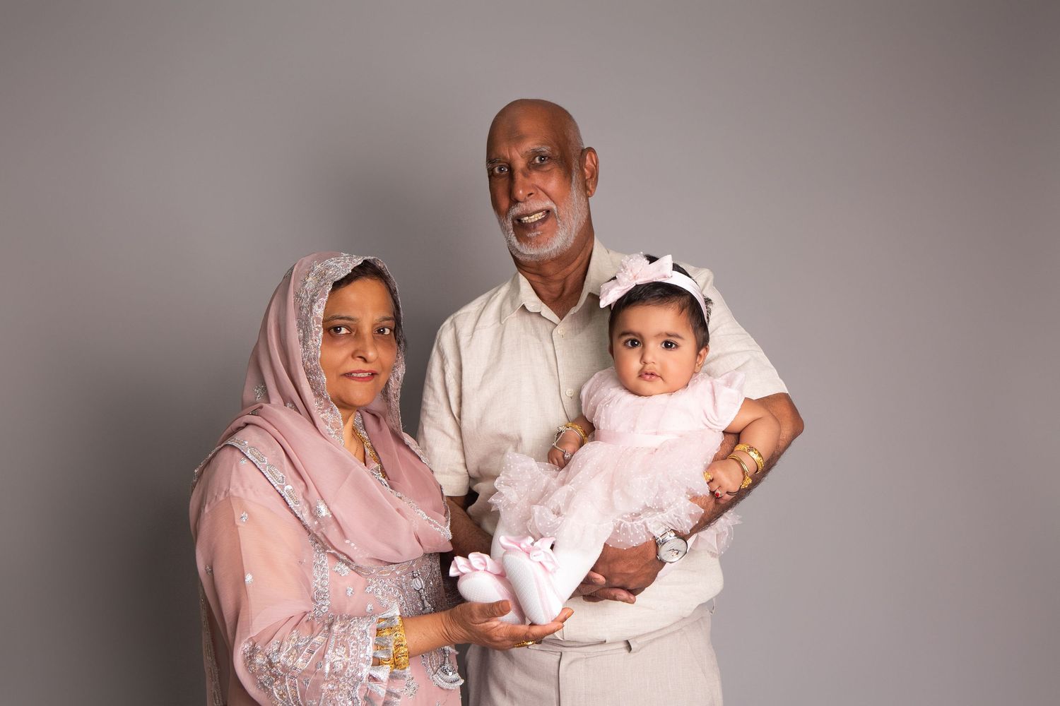 Elderly South Asian couple in traditional attire smiling, grandfather holding a baby girl in pink dress against gray background.
