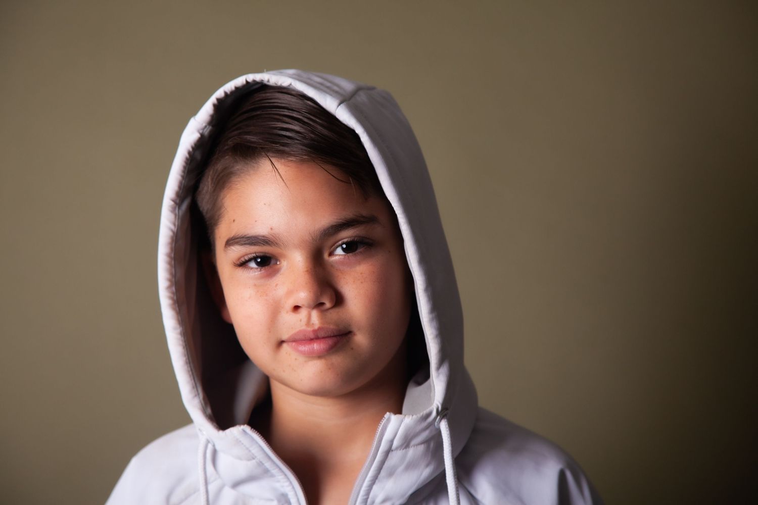 Young girl in white hoodie posing confidently for portrait against neutral olive background indoors.