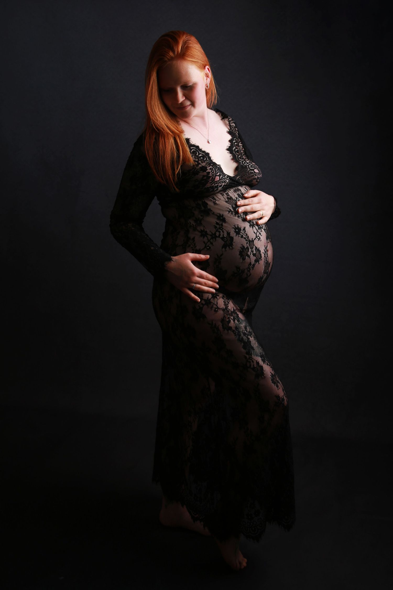 Pregnant redhead woman in sheer black lace gown posing in dramatic studio lighting.