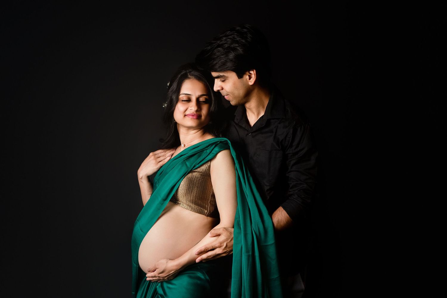 Pregnant Indian woman in green saree with partner embracing her from behind on dark background.