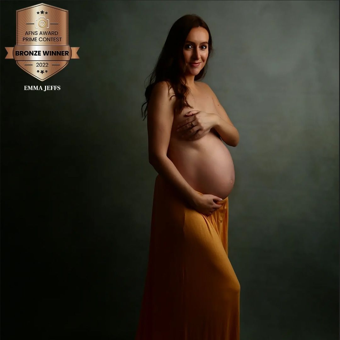 A pregnant woman in a golden yellow skirt poses in an award-winning bronze maternity studio portrait by Emma Jeffs.