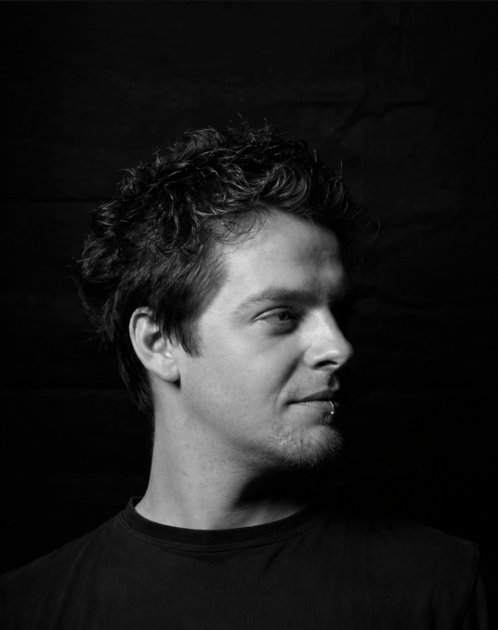 Black and white profile portrait of a young man with curly hair and a chin piercing wearing a dark shirt.