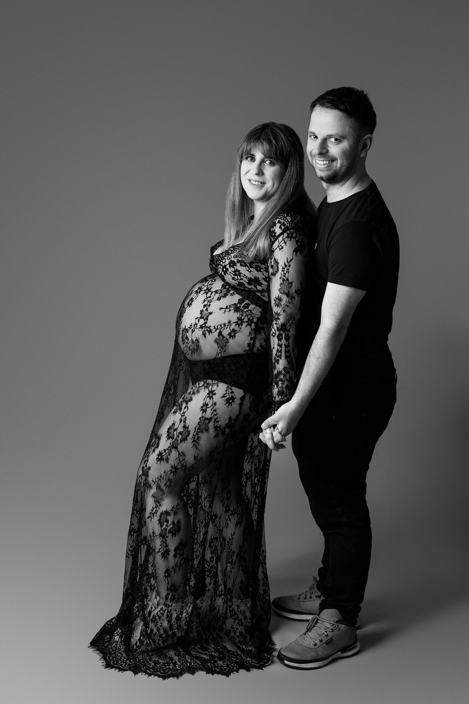 Black and white full-length maternity portrait of couple holding hands, woman in elegant lace gown.