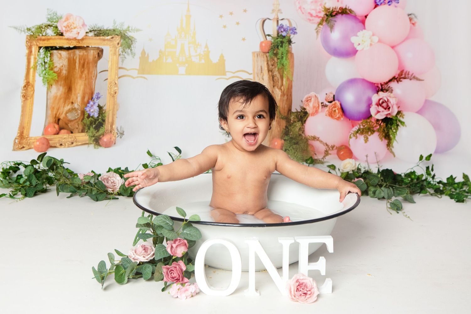 Baby sitting in a white tub surrounded by balloons and flowers at a first birthday cake smash photo shoot.