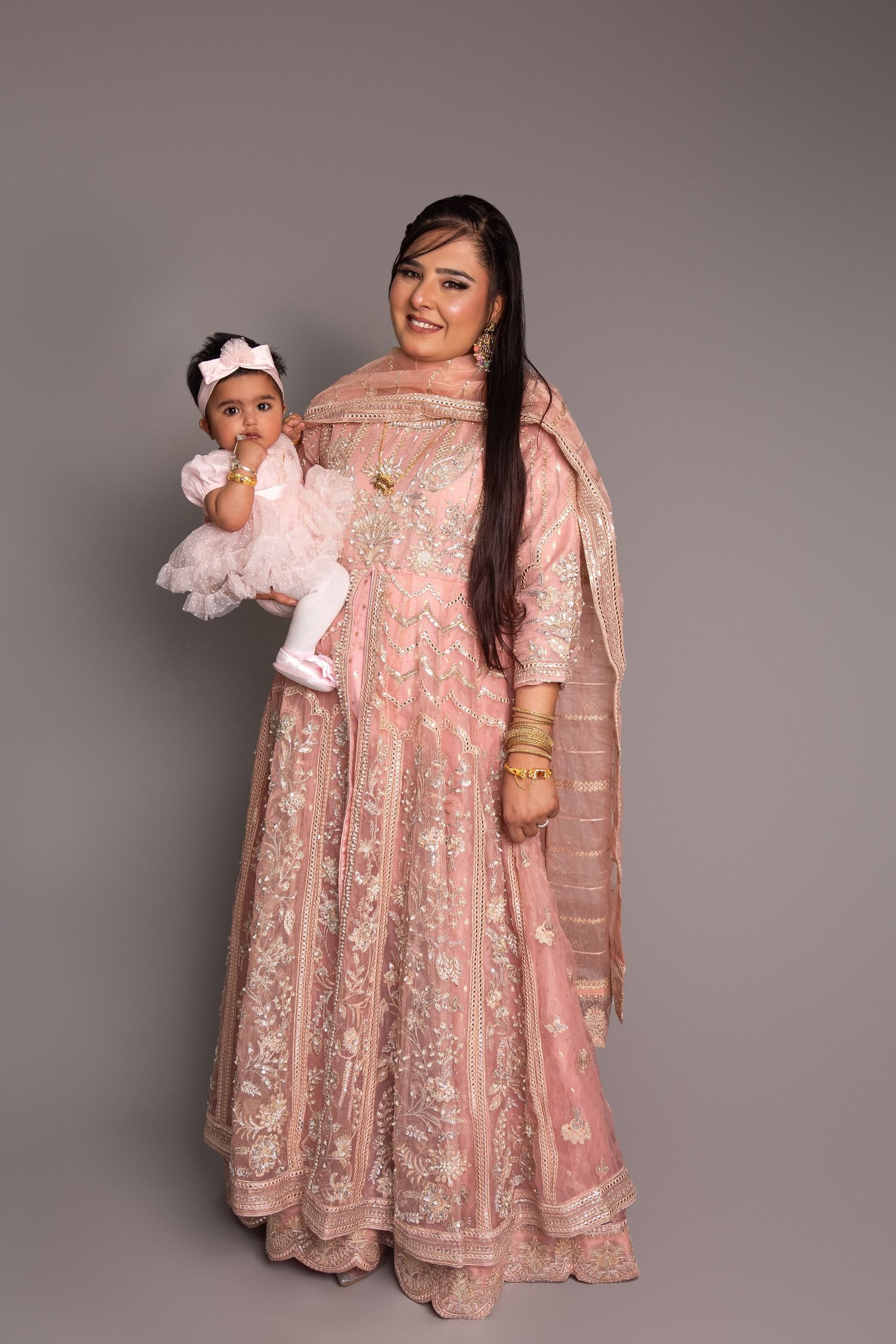 A woman in a pink embroidered anarkali suit holds a baby girl in a white dress against a gray background.