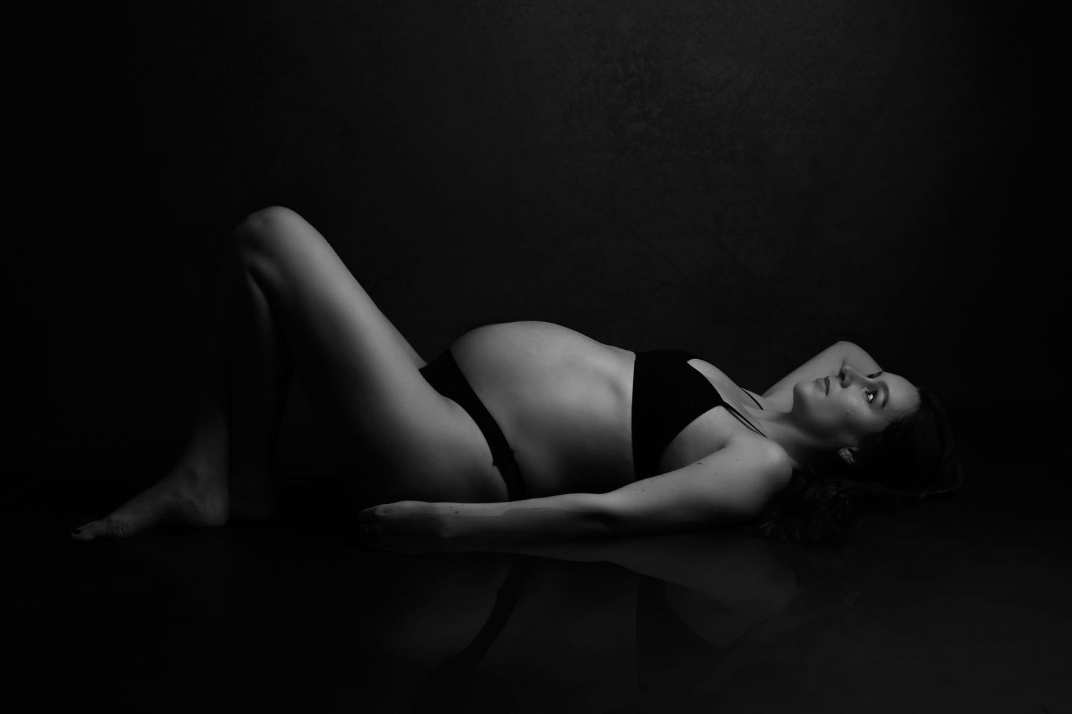 Black and white maternity photo of pregnant woman in lingerie lying gracefully on studio floor.