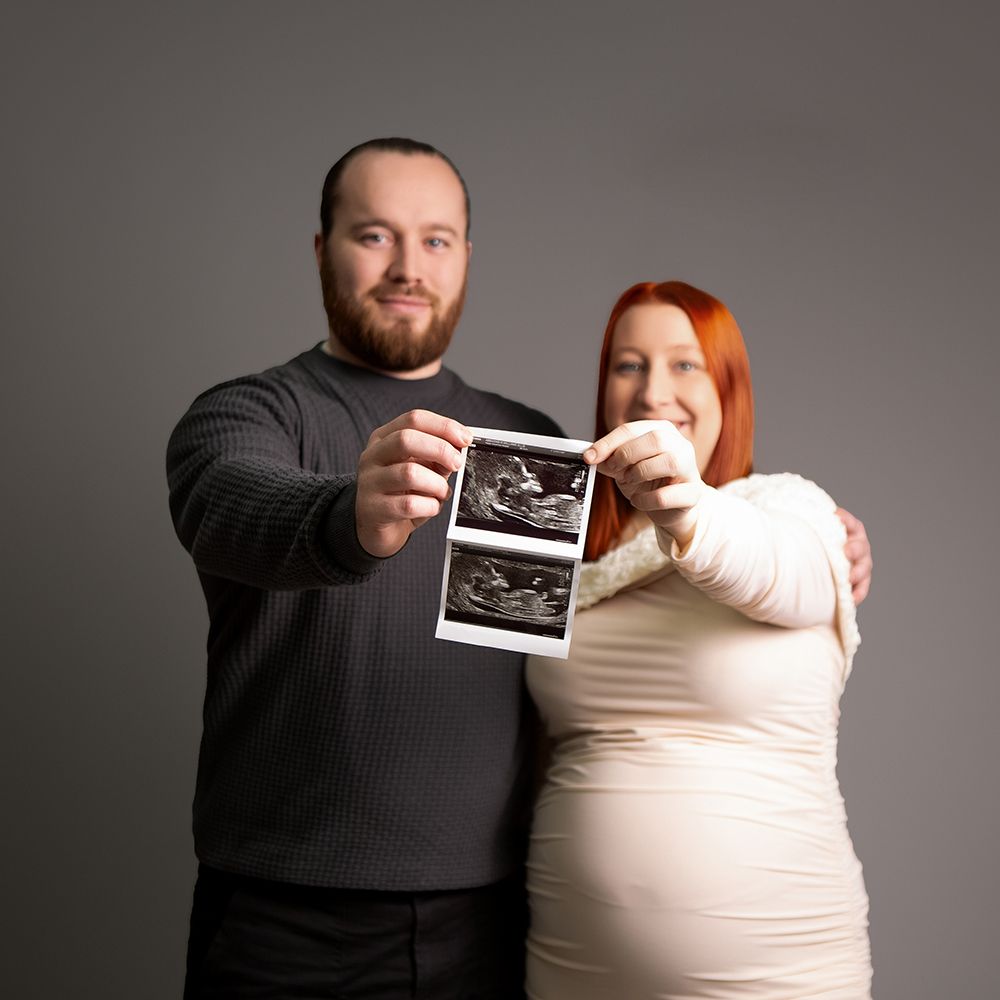 Pregnant couple smiling and holding ultrasound photos together against a gray studio background.