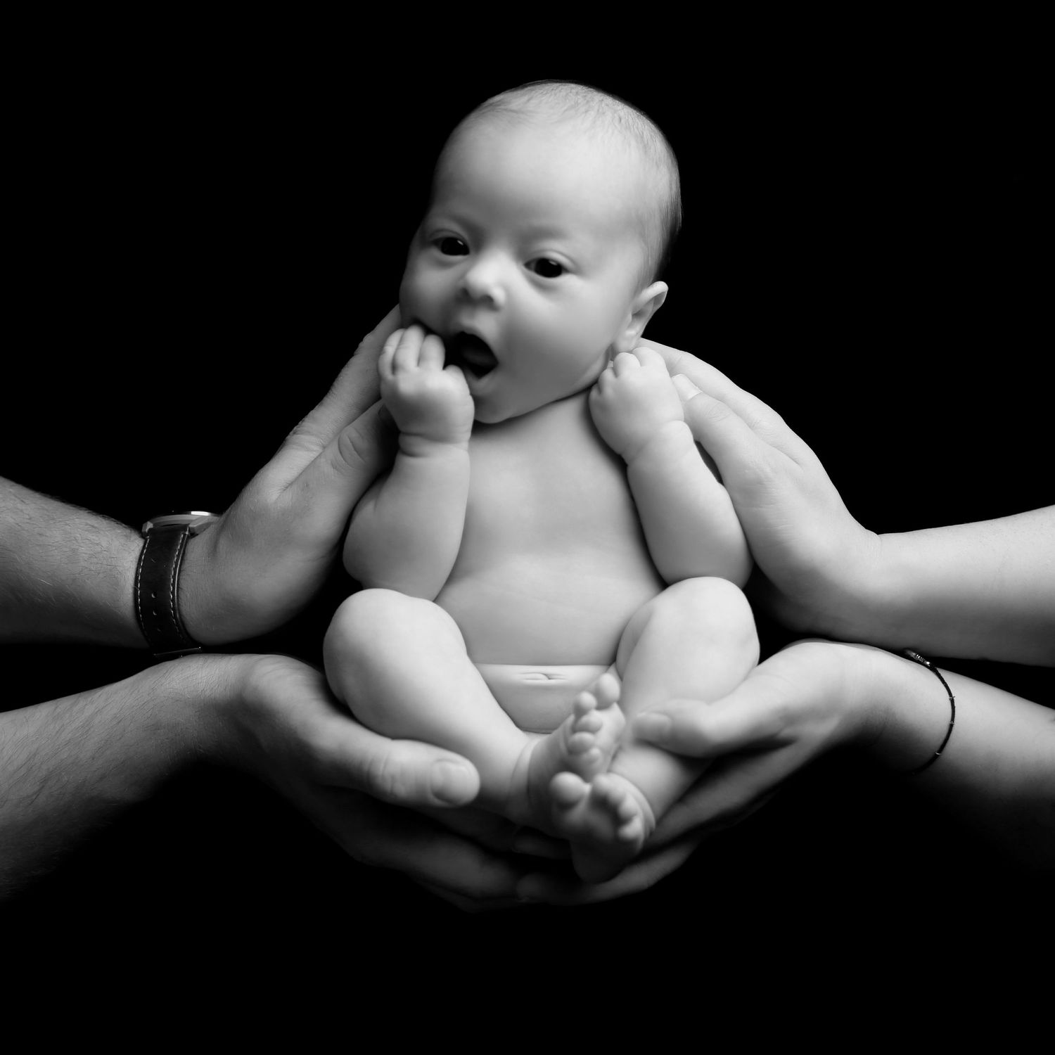 Black and white photo of a newborn baby cradled in the hands of two adults against a dark background.