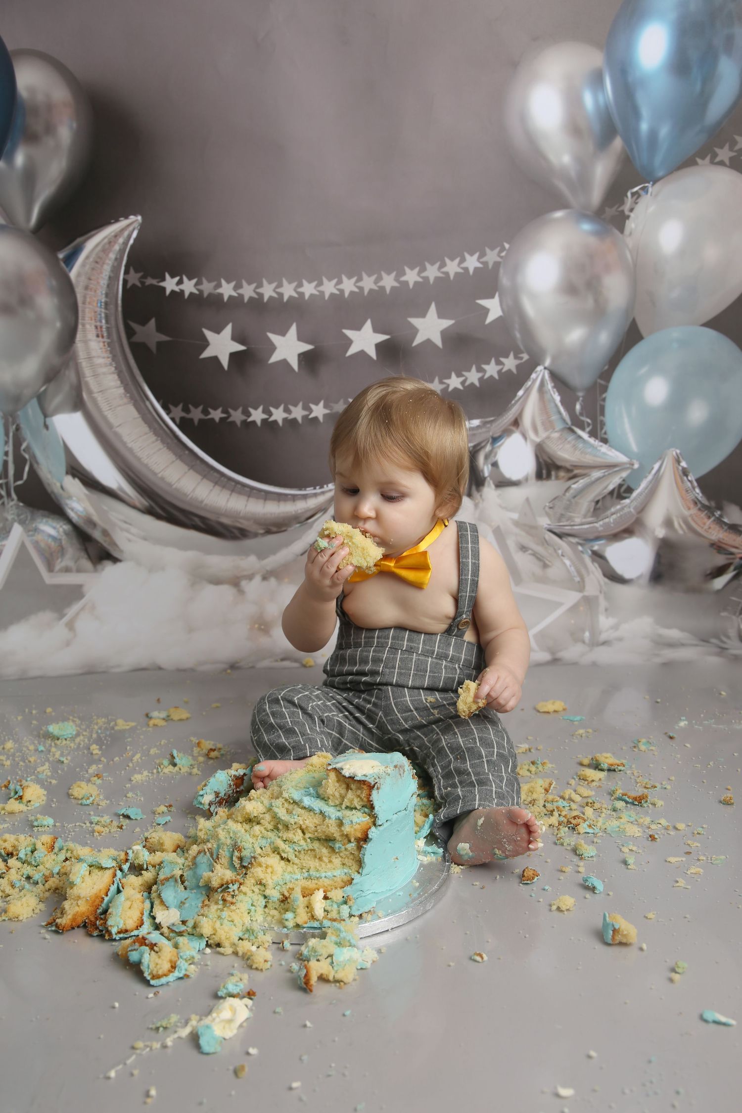 Baby boy in suspenders smashes first birthday cake surrounded by blue balloons and moon decorations.
