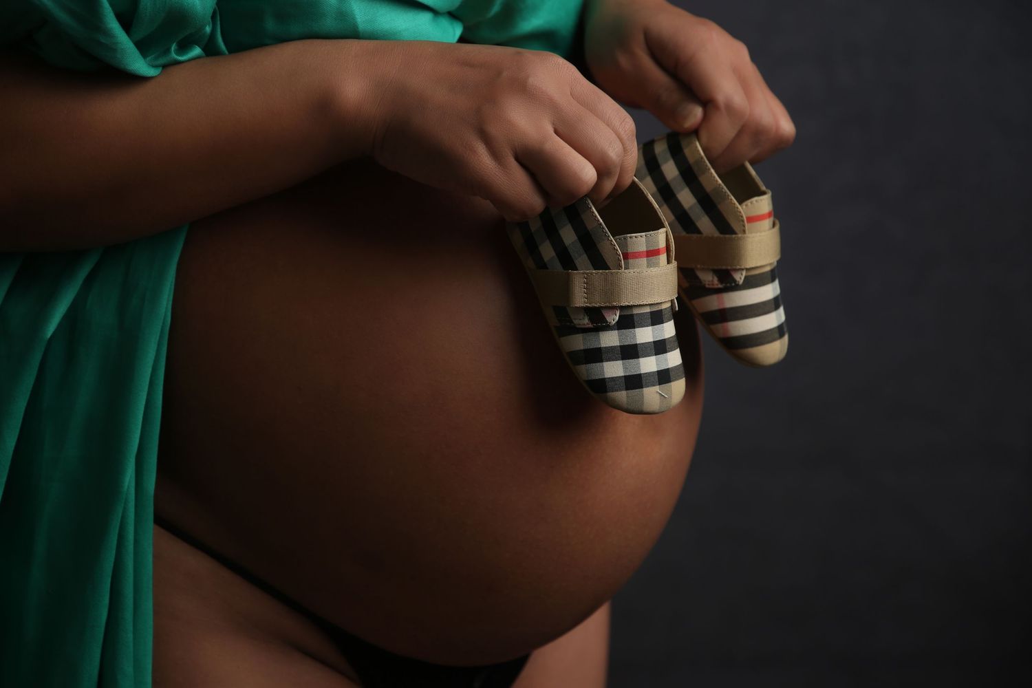 Pregnant woman in green robe holding tiny Burberry plaid baby shoes against her bare belly.