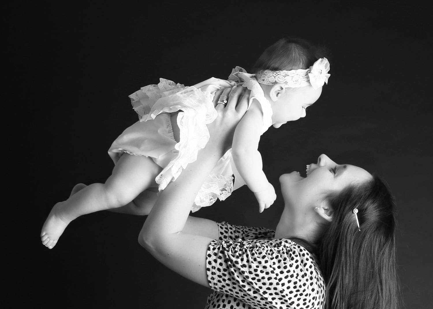 Black and white photo of a joyful mother lifting her baby girl in a white dress above her head.