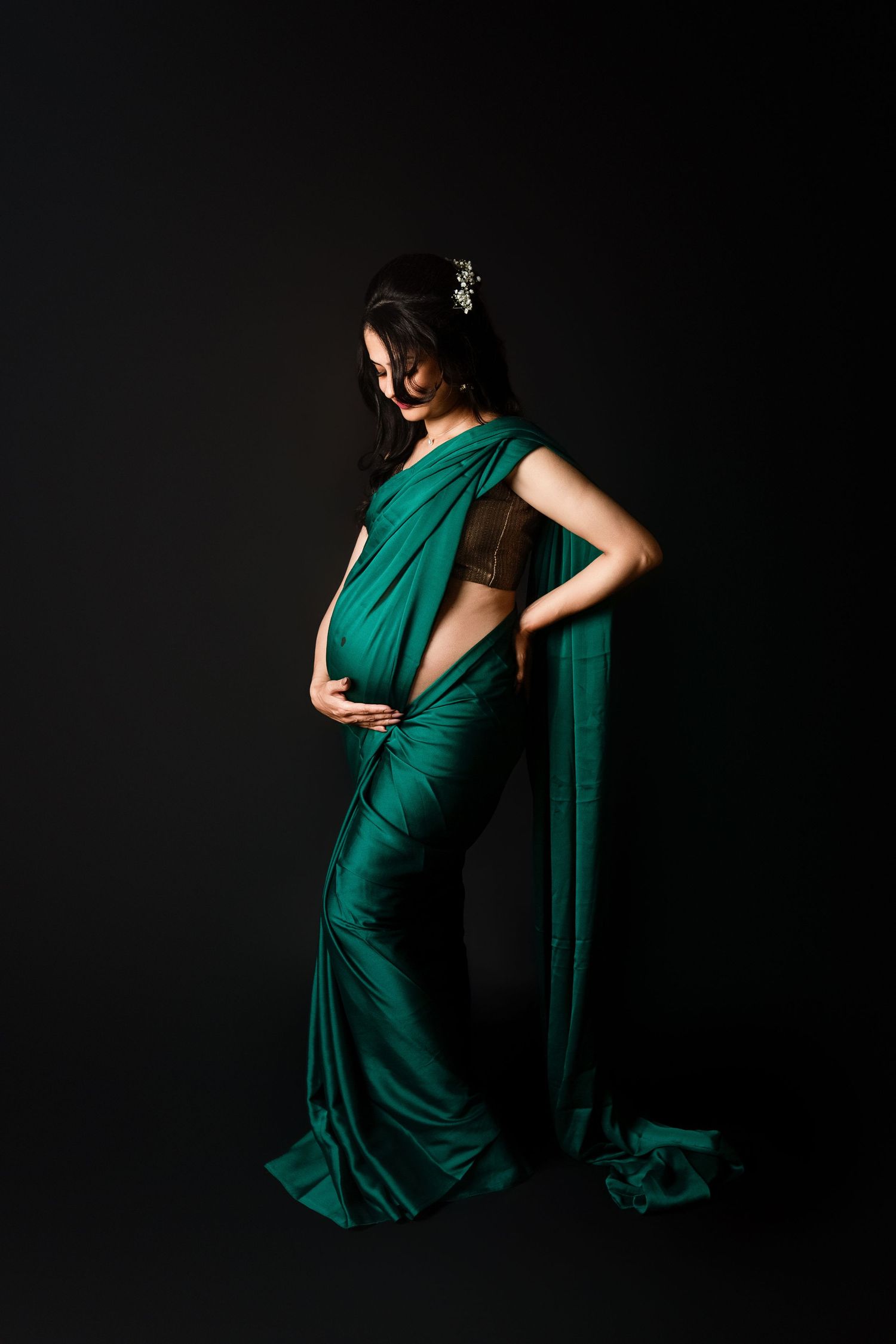 Elegant pregnant woman in emerald green silk saree with gold blouse posing on black background.