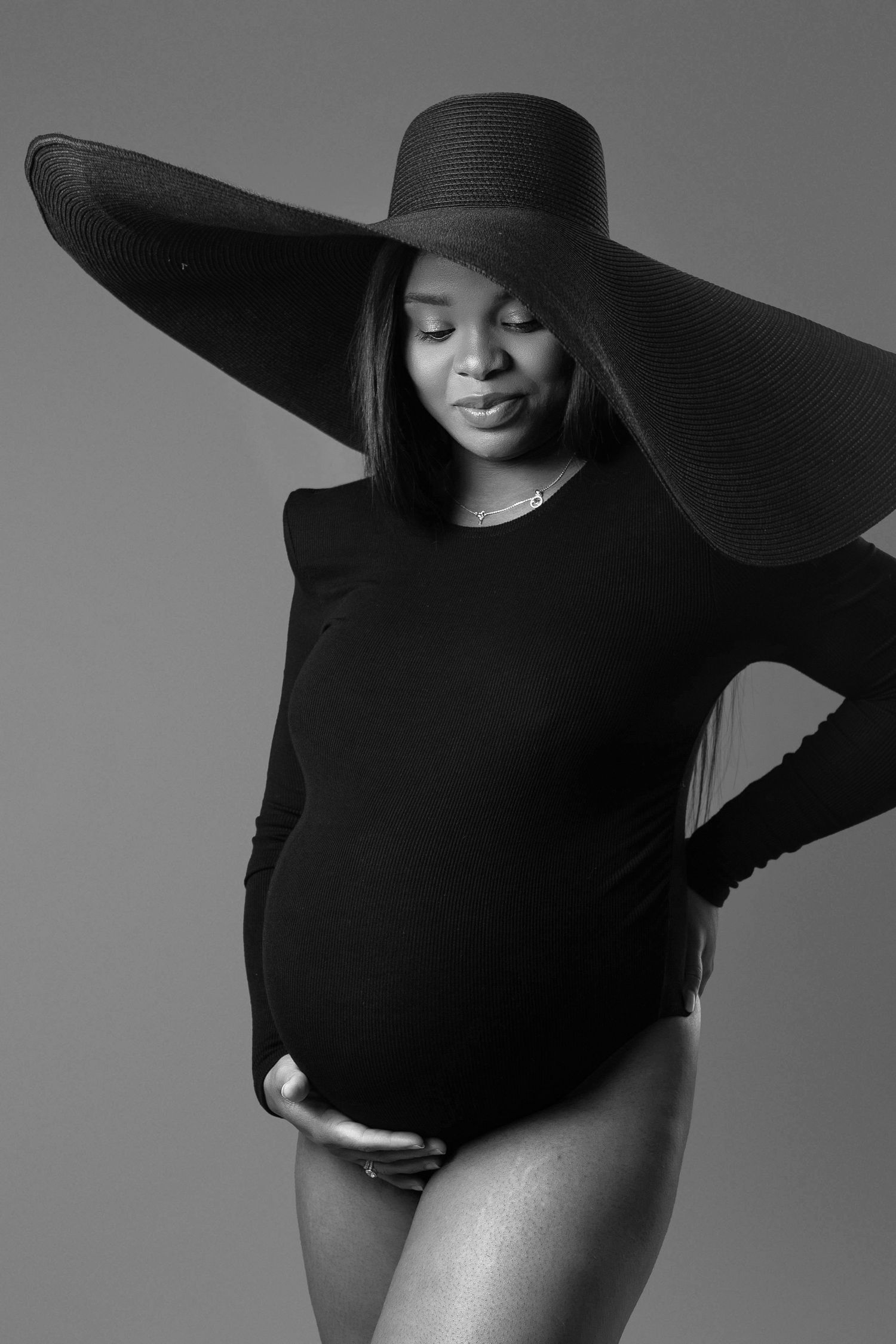 Black and white maternity portrait of pregnant woman in black bodysuit and wide hat.