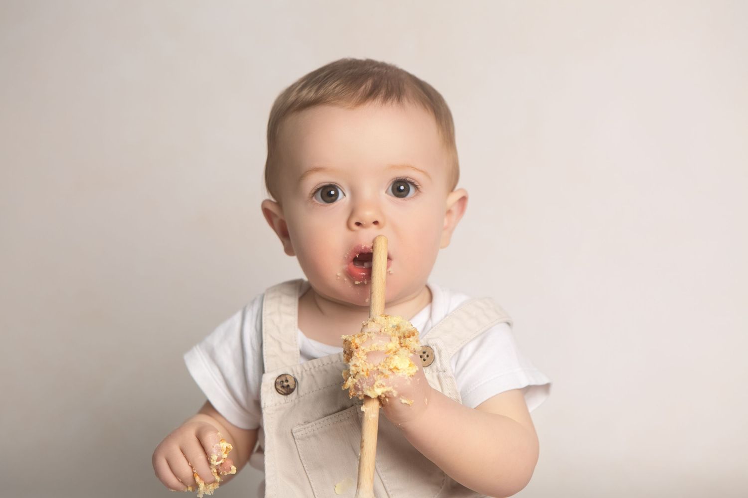 Adorable baby boy in suspenders chewing on a decorative gold star wand against a light gray background.