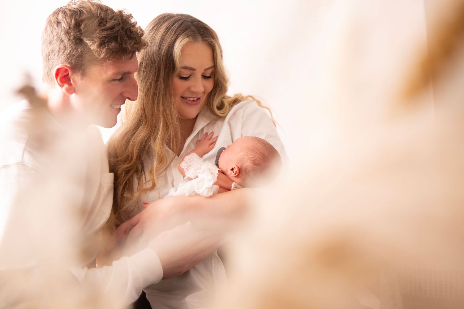 Young couple smiling and bonding with their newborn baby in a bright, warmly lit indoor setting.