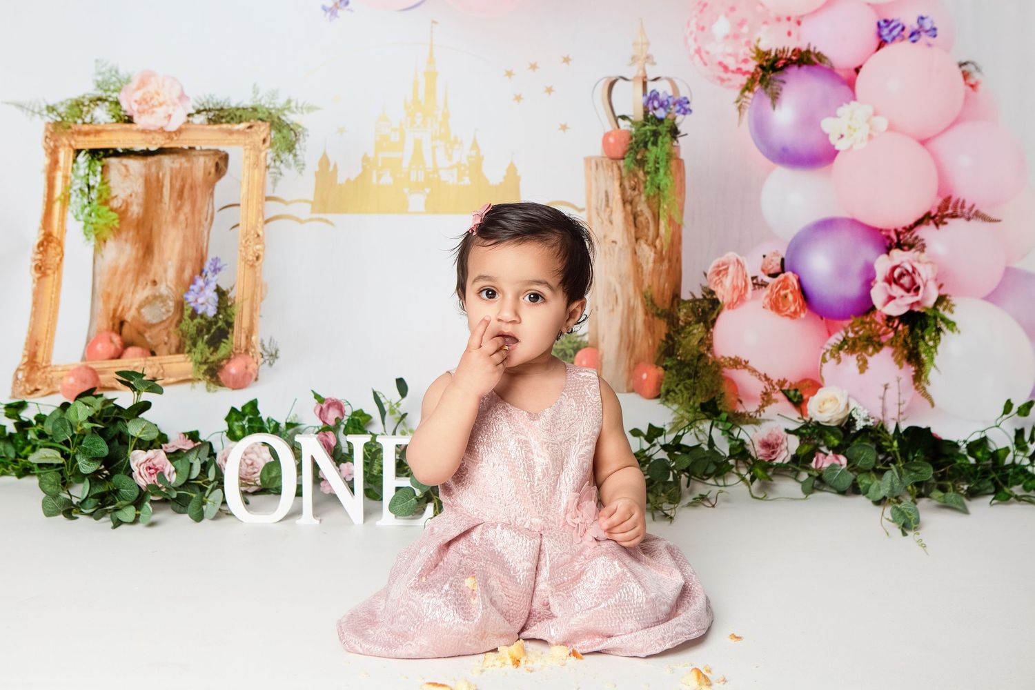 Baby girl in pink dress celebrates first birthday with ONE sign, floral decor, and pastel balloons.