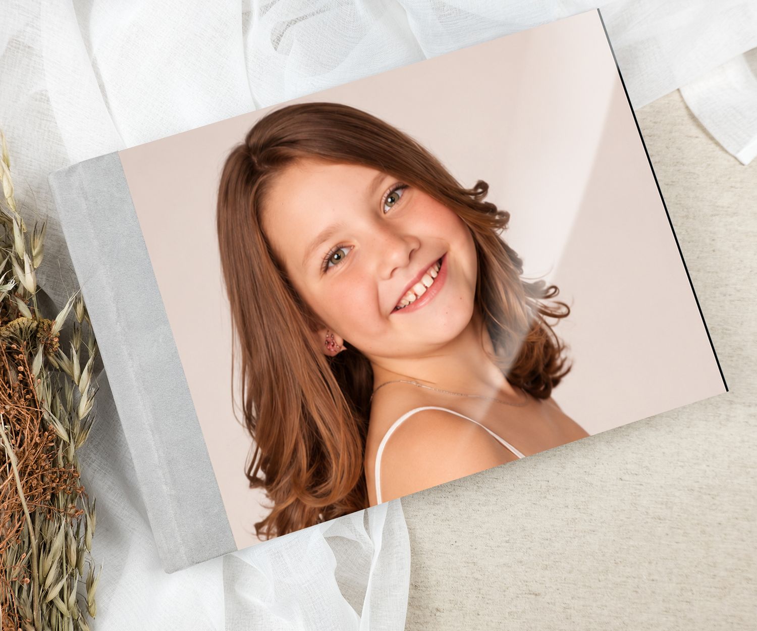 A smiling young girl with brown hair featured on a photo book cover, surrounded by soft fabric and dried flowers.