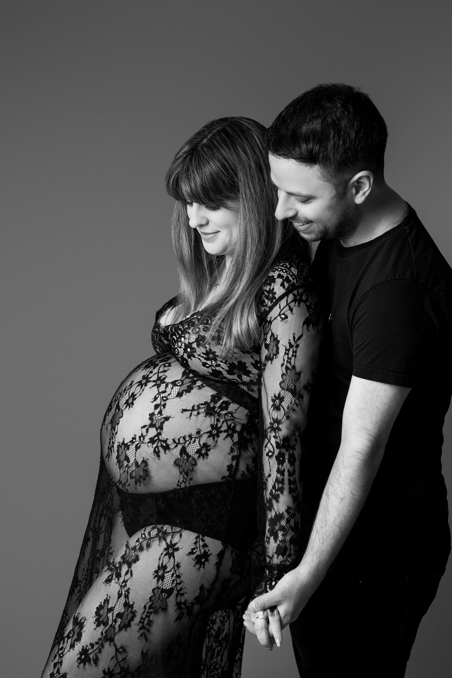 Black and white studio photo of couple, man gazing at pregnant partner in sheer floral lace dress.