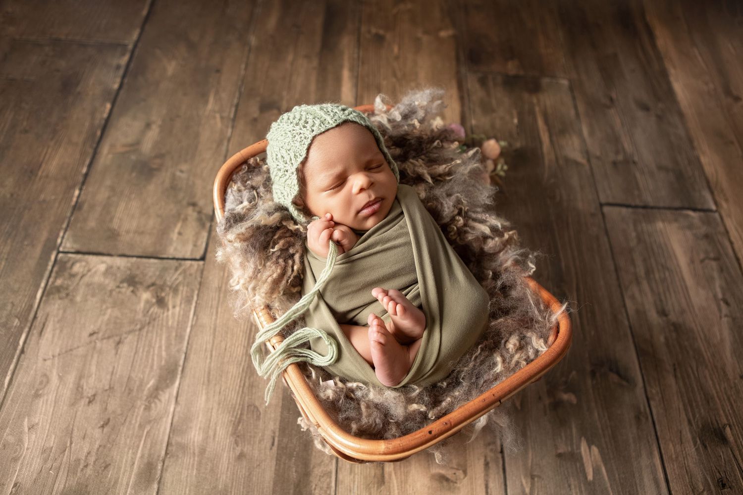 Newborn baby wrapped in green swaddle with bonnet sleeping in a wicker basket lined with fluffy brown fur.