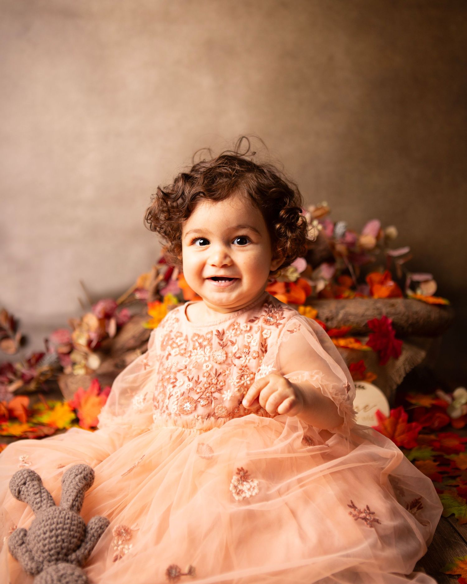 Smiling baby girl in a pink tulle dress surrounded by autumn leaves and flowers in a studio portrait.