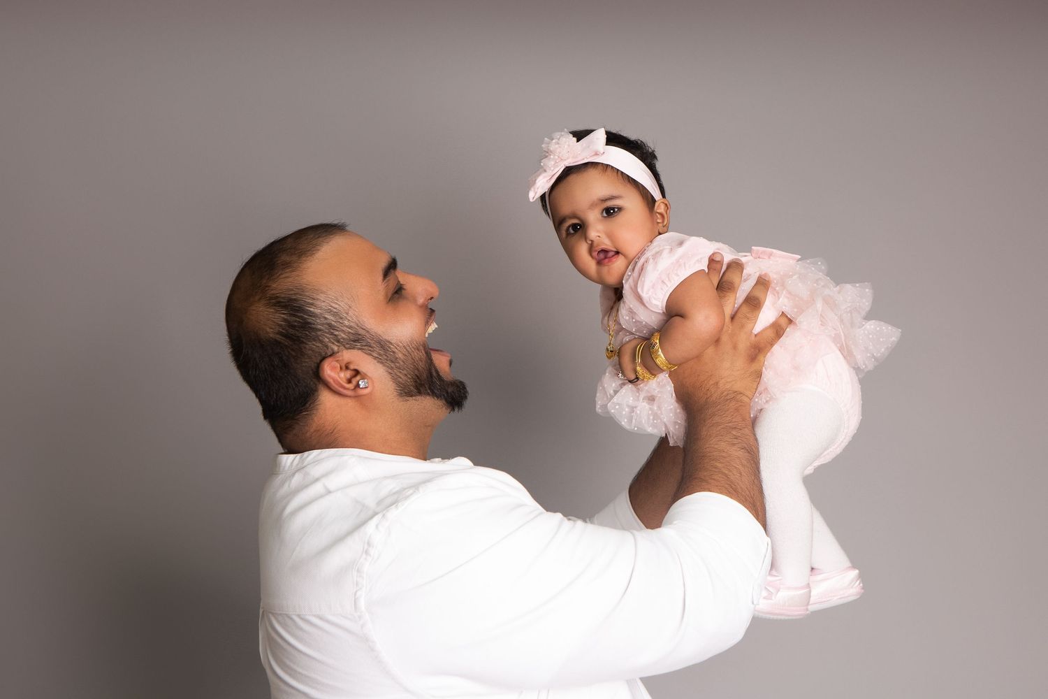 A father in a white shirt joyfully lifts his smiling baby girl in a pink tutu dress against a gray background.