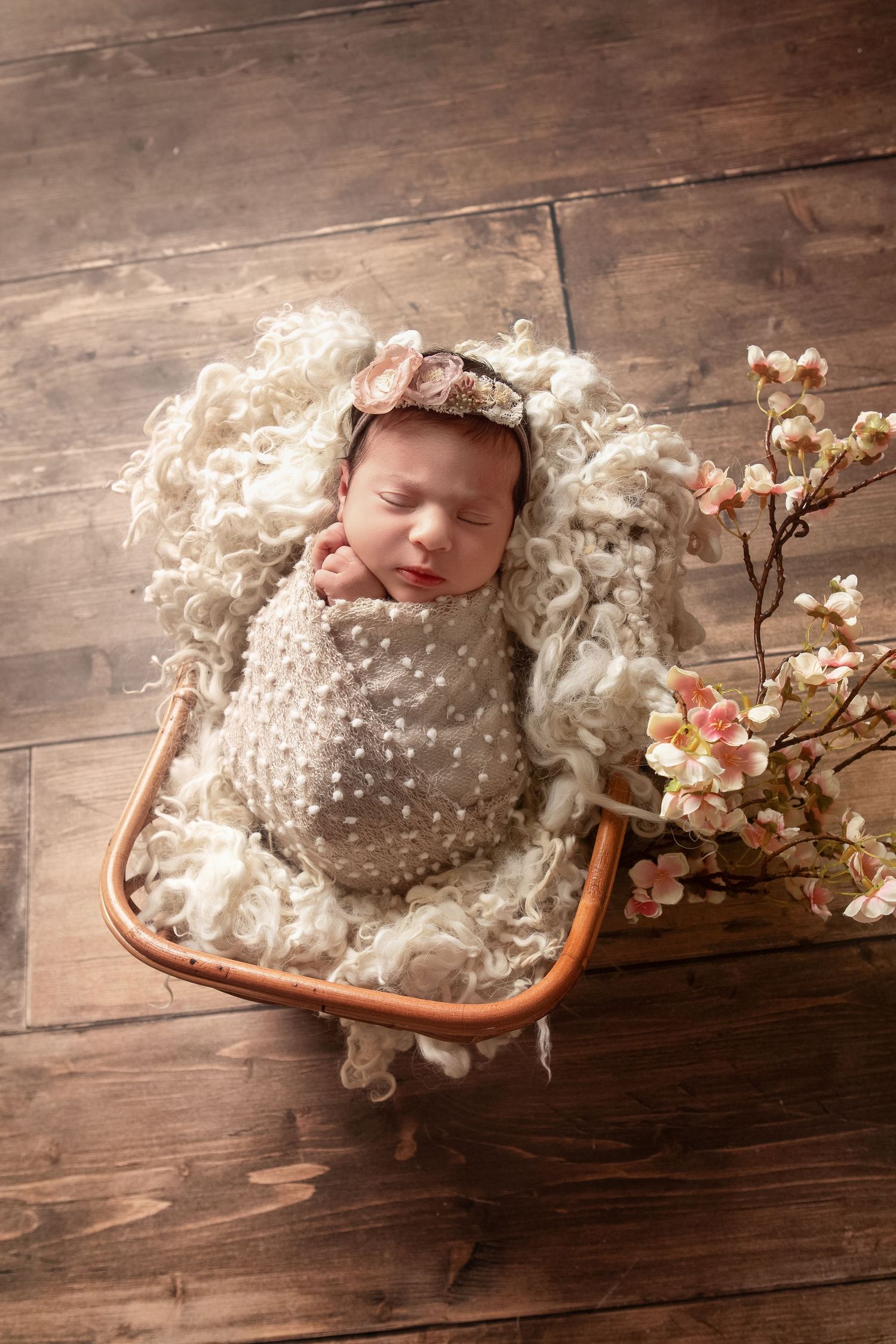 Newborn baby swaddled in beige wrap sleeping in a basket with fluffy wool lining and pink cherry blossoms on wood floor.
