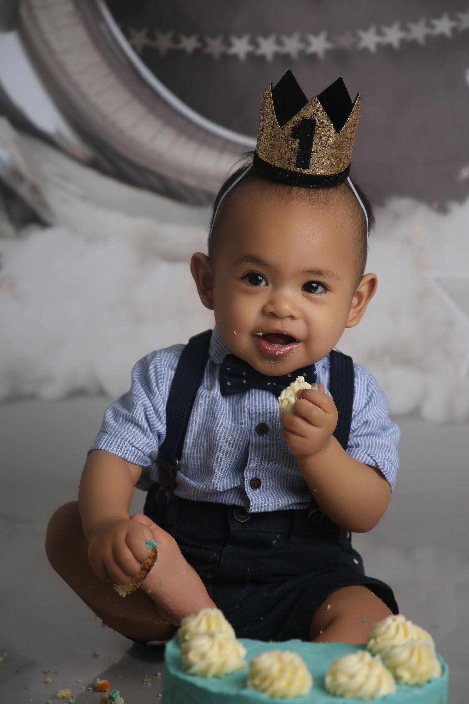 A baby boy in suspenders and bow tie wears a gold crown headband while eating his first birthday cake.