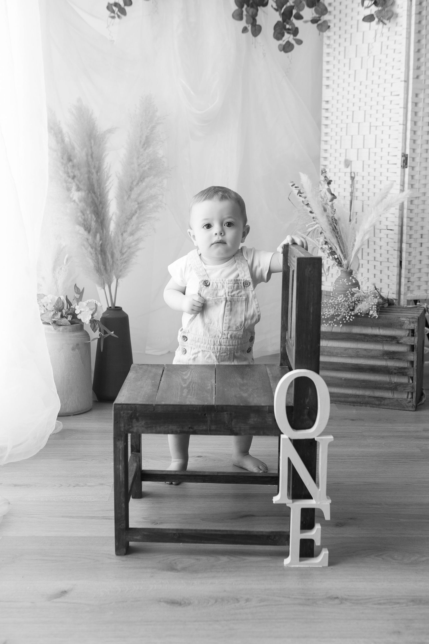 Black and white photo of a baby standing at a wooden table with ONE sign, first birthday portrait.