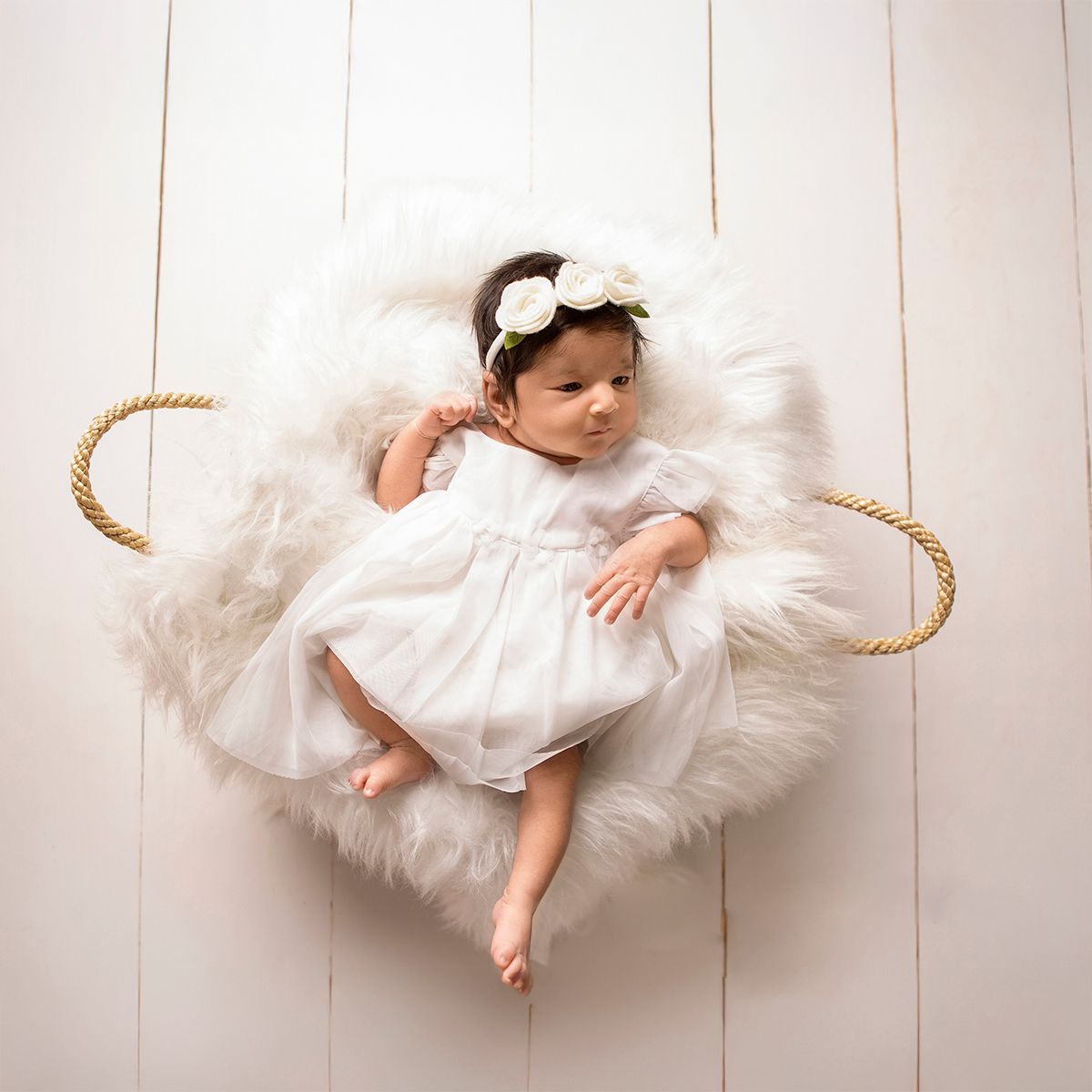 Newborn baby girl in white dress and floral headband lying in a fur-lined basket for a professional photo session.