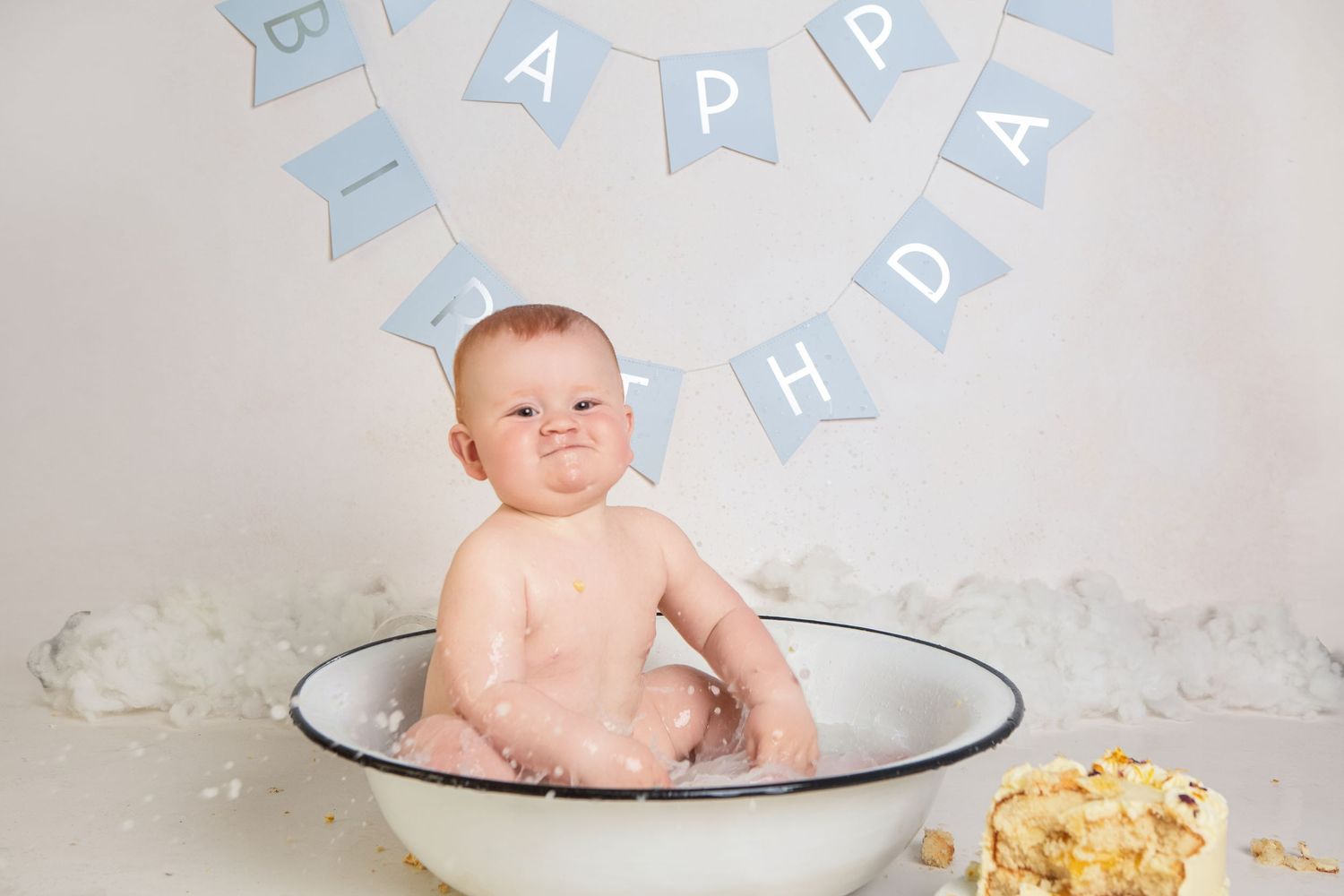 Baby sitting in a white enamel tub during a first birthday cake smash photo session with a Happy Birthday banner.