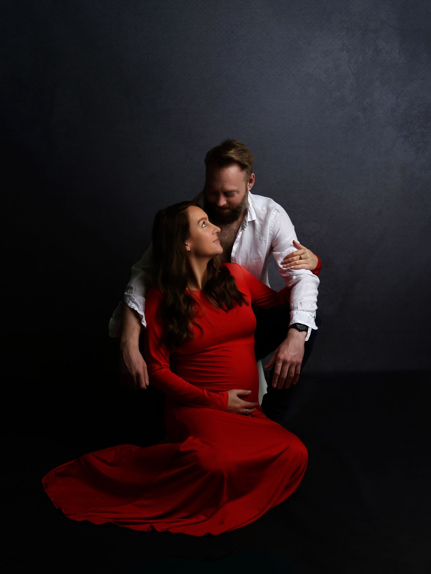 A woman in a red dress sits on the floor gazing up at a man in a white shirt in a dramatic studio portrait.